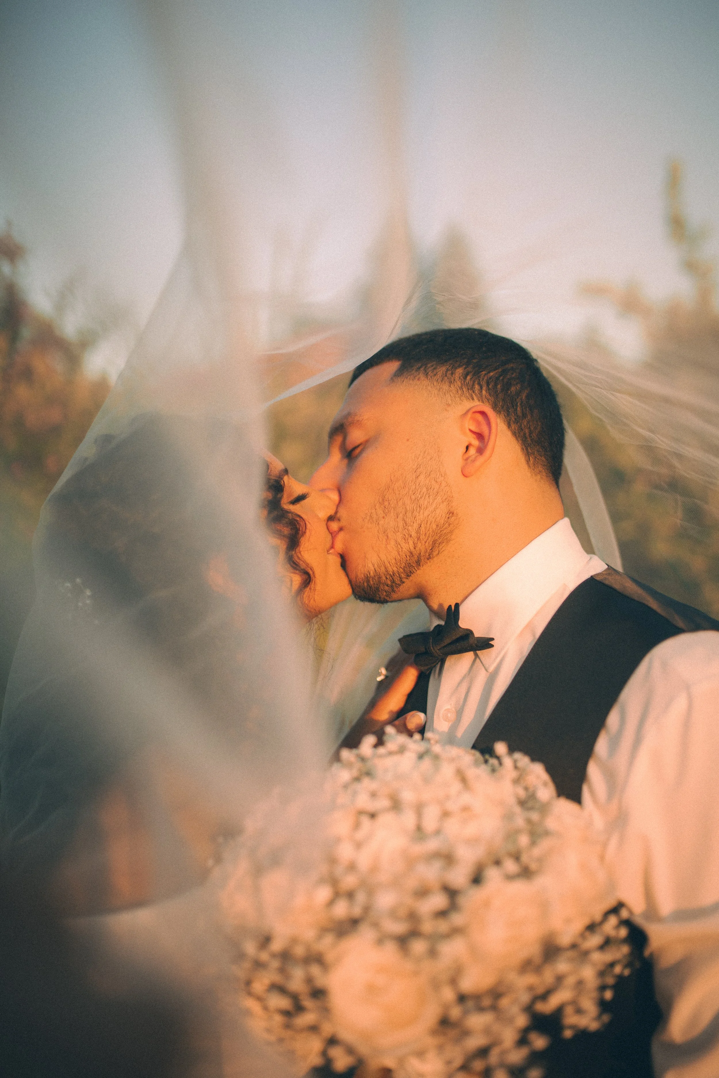 A couple sharing a kiss underneath a wedding veil at sunset, with the bride's bouquet of flowers in the foreground.