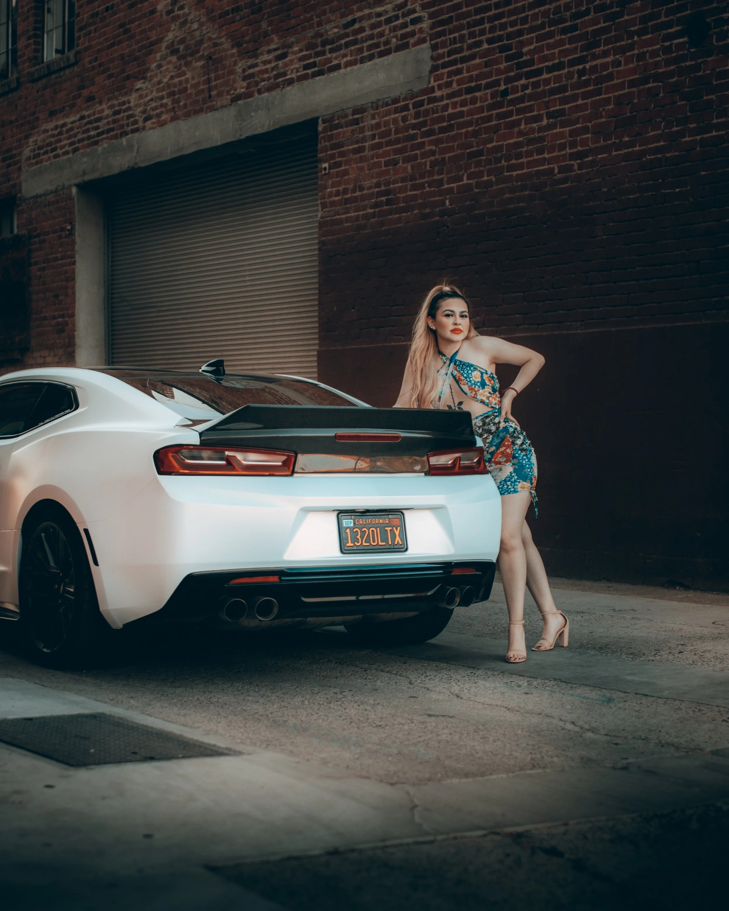 A woman with long blonde hair, wearing a colorful floral dress and beige high heels, stands beside a white sports car with a black roof and orange California license plate, in an urban alley with a brick wall and a rolled-up garage door.