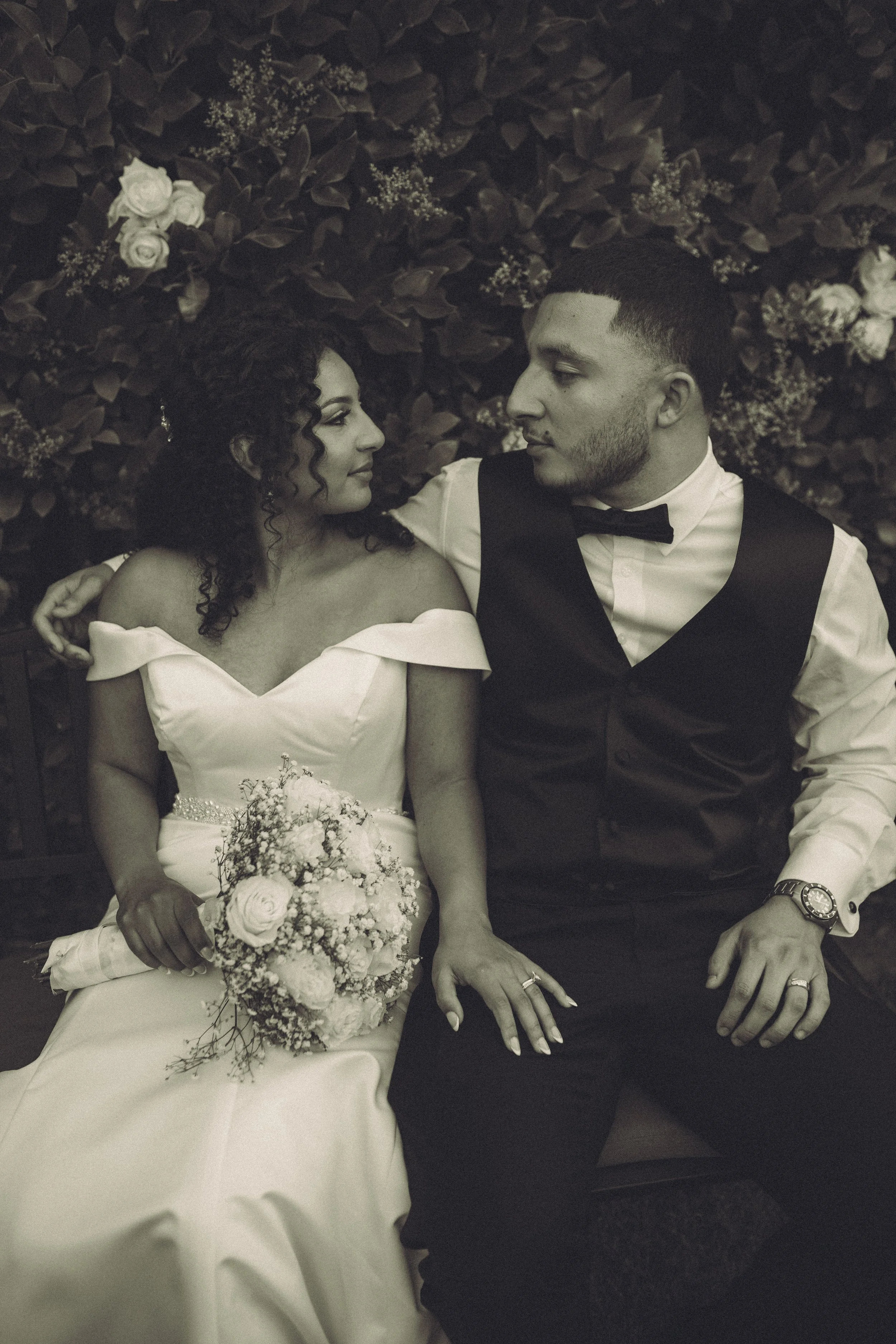 A black-and-white photo of a bride and groom gazing at each other, seated against a background of dark leafy plants and white flowers. The bride is holding a bouquet and the groom is wearing a suit with a bow tie.
