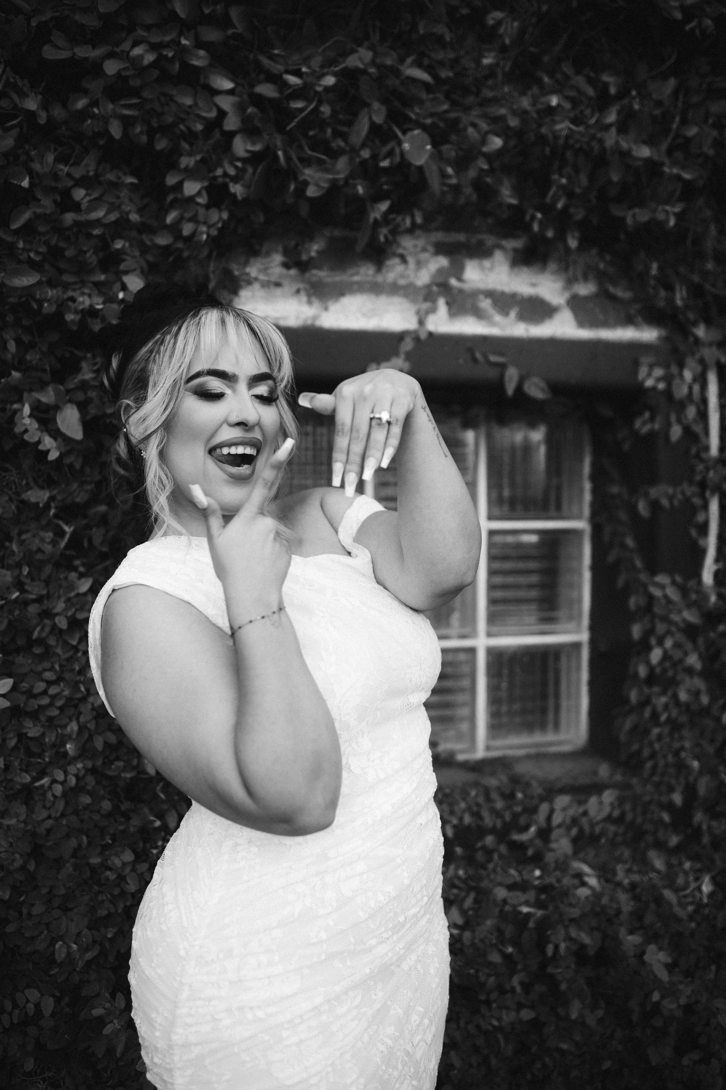 A woman in a white lace dress poses with a playful expression, holding her hand near her face, outdoors with leafy bushes and a small building in the background.