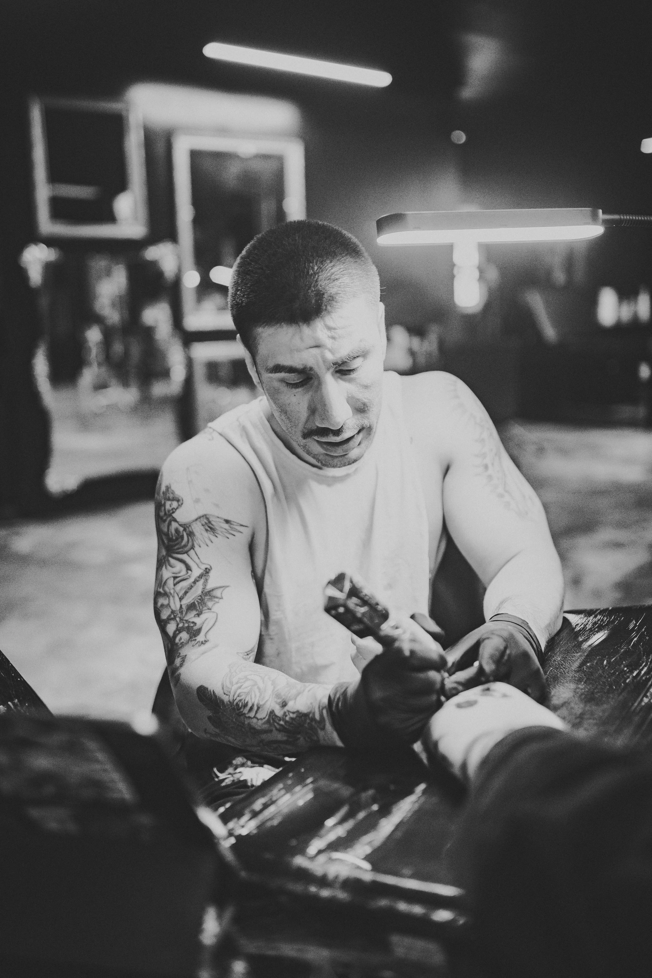 Black and white photo of a tattooed man in sleeveless shirt, leaning forward, looking at his phone, with a tattoo sleeve on his left arm, in a dimly lit room.