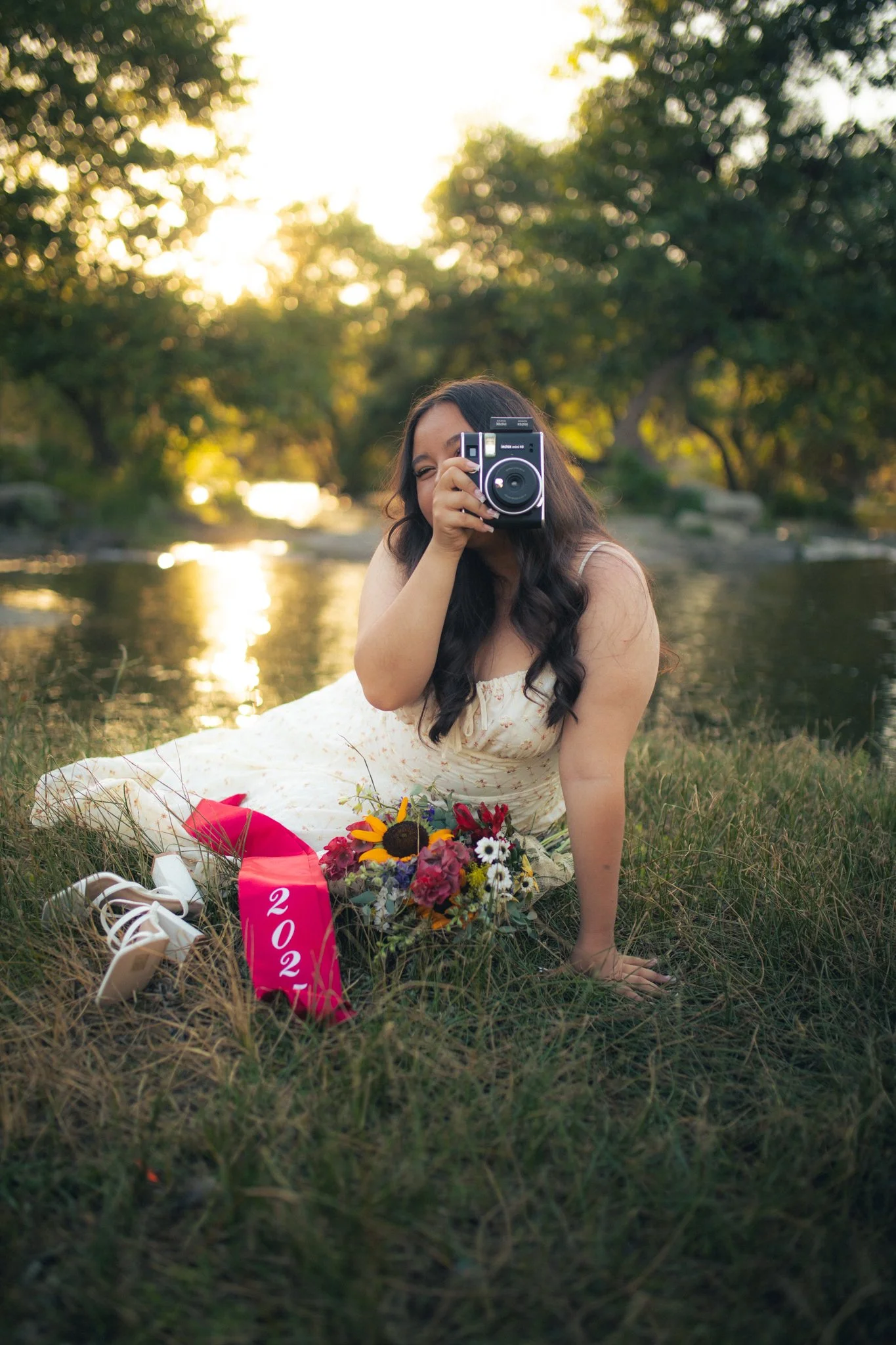 A woman with long dark hair sitting on grass near a river, holding a vintage camera up to her face, with a bouquet of flowers, a pink ribbon that says '2023,' and white sneakers nearby, during sunset with trees in the background.
