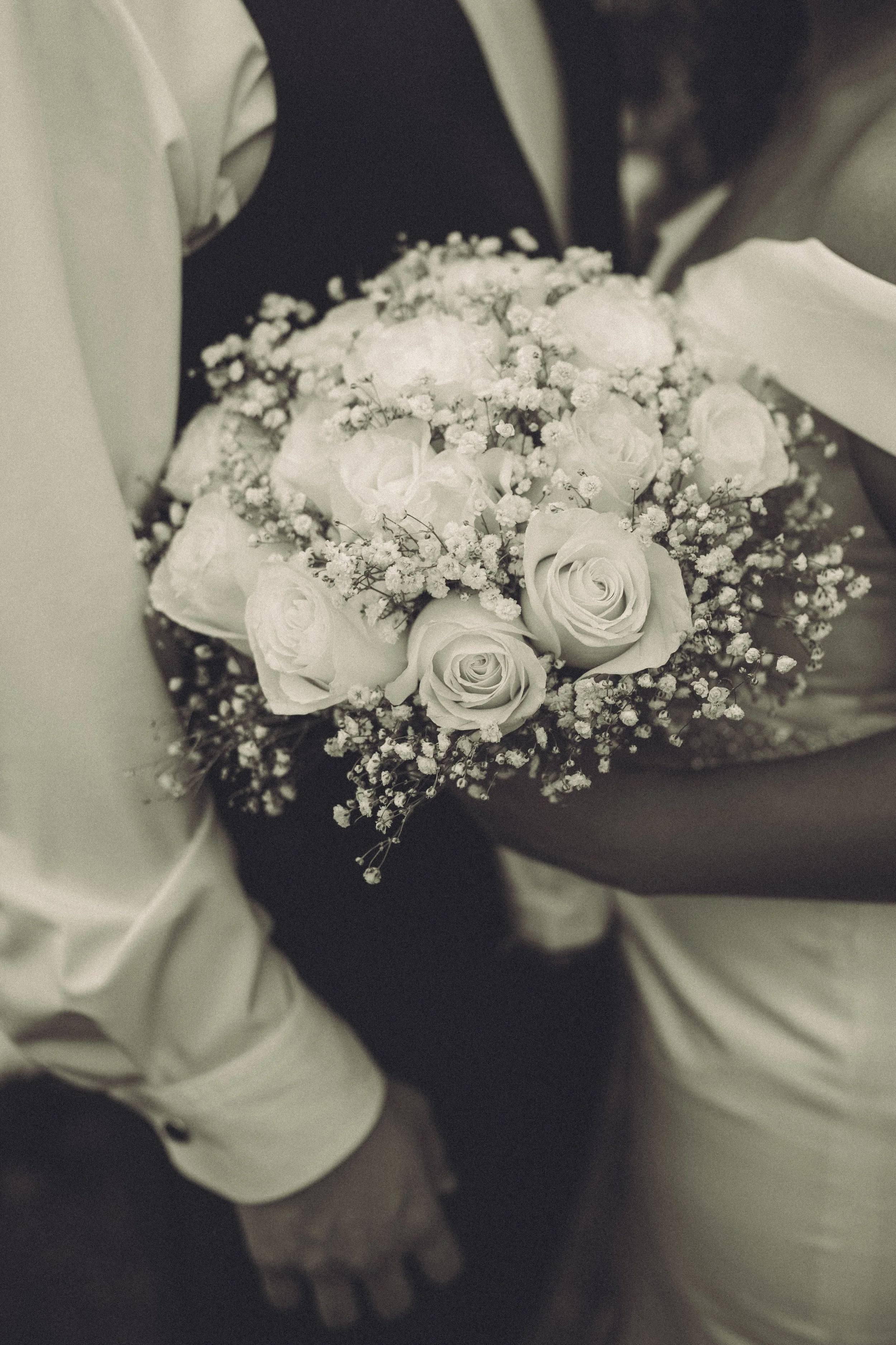 A bouquet of white roses, baby's breath, and other white flowers being held by someone wearing a white shirt and black pants.