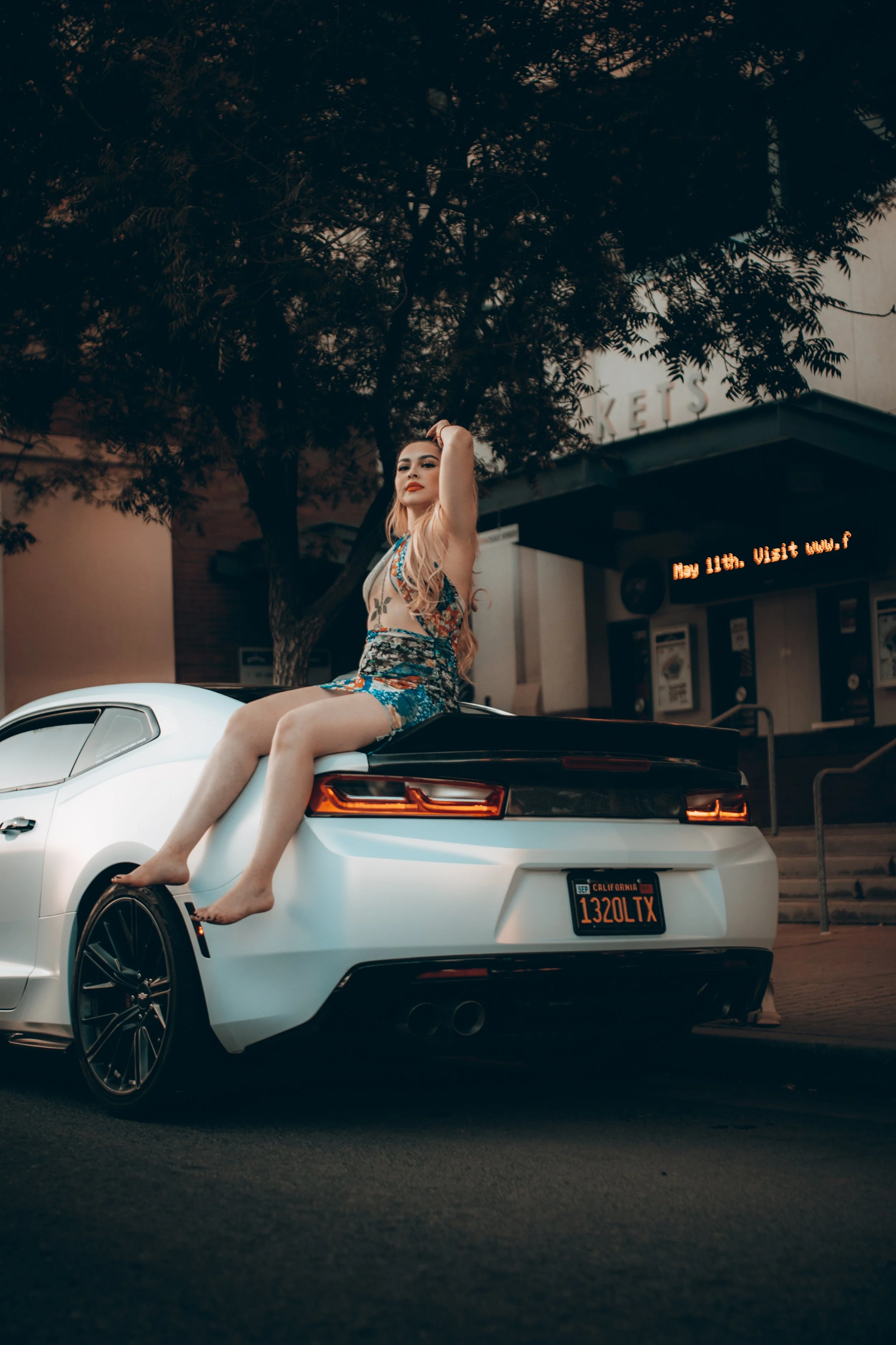 A woman with long blonde hair and red lipstick sitting on the trunk of a white sports car with California license plates, parked outdoors in front of a building with a sign that says 'KET'S' and trees overhead.