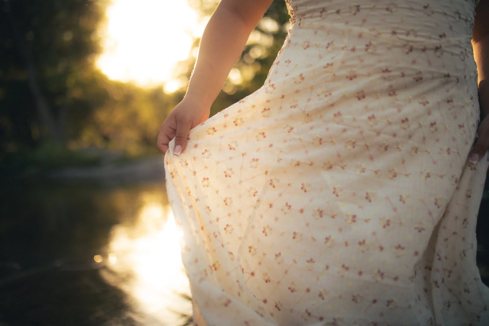 Close-up of a woman holding the hem of her floral dress outdoors at sunset, with trees and sunlight in the background.