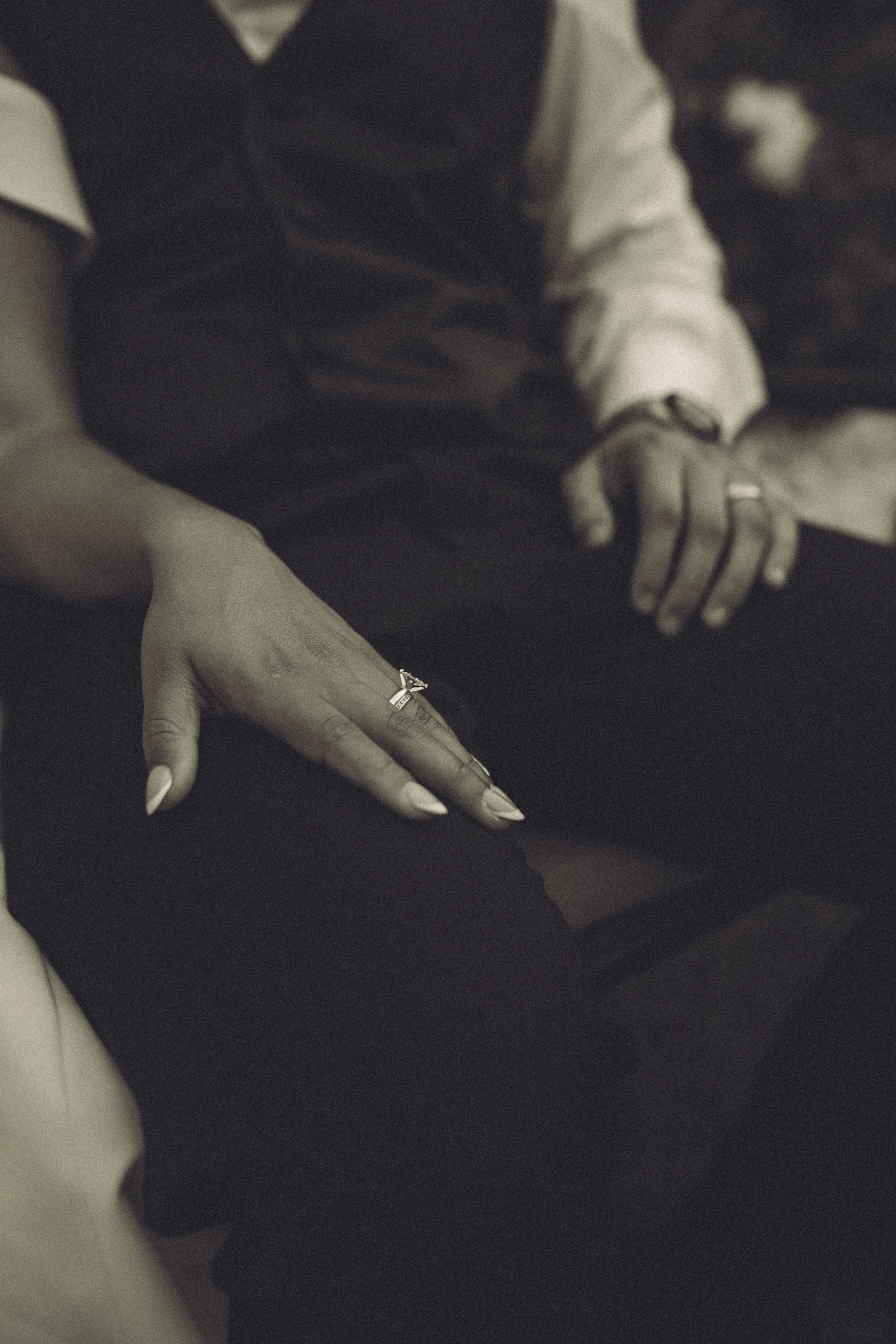 Close-up of two hands, one with long painted nails and the other with rings, resting on dark fabric, with a person in a blazer and shirt in the background.