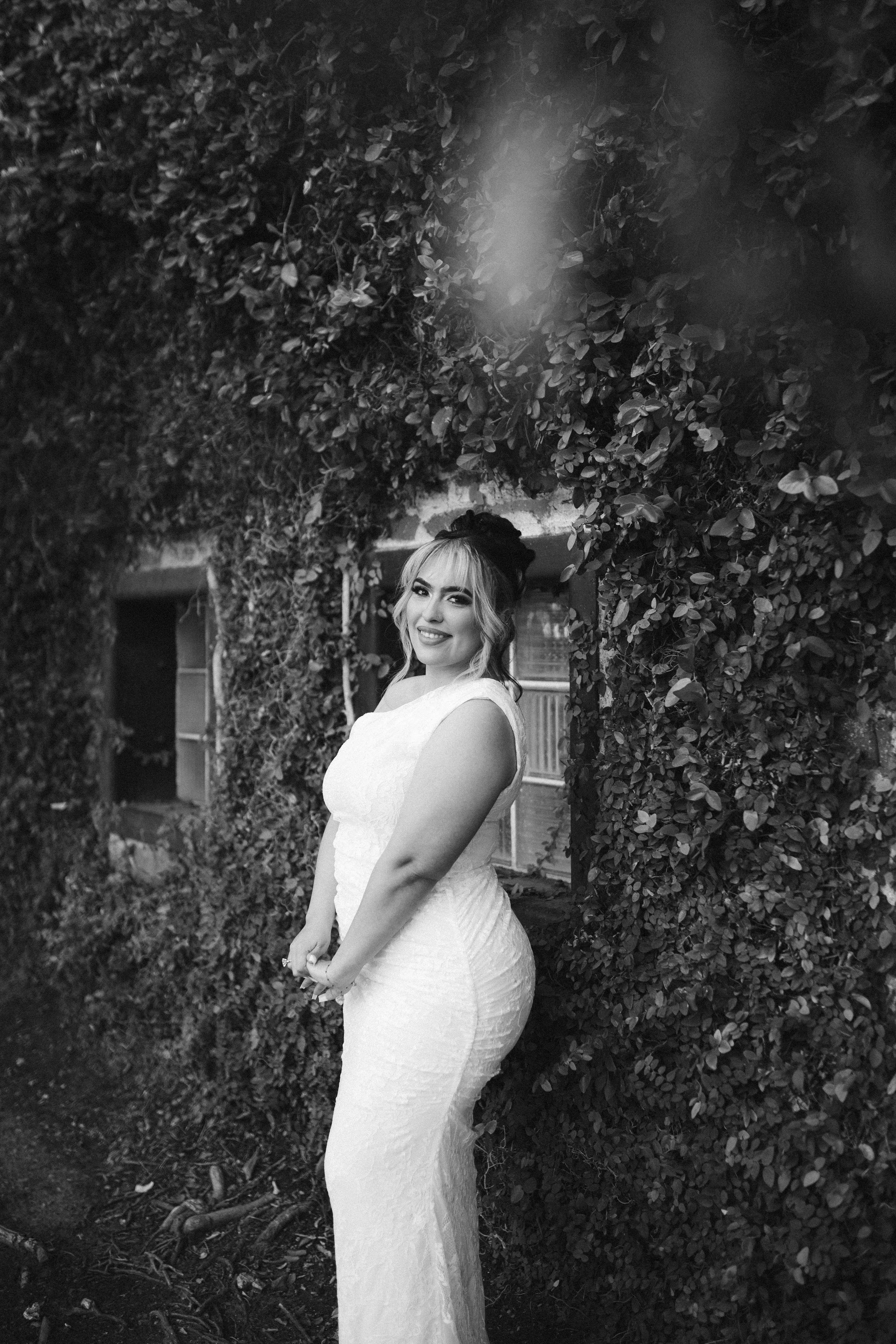 A woman in a white dress standing outdoors near a bush with small leaves, smiling and looking at the camera.
