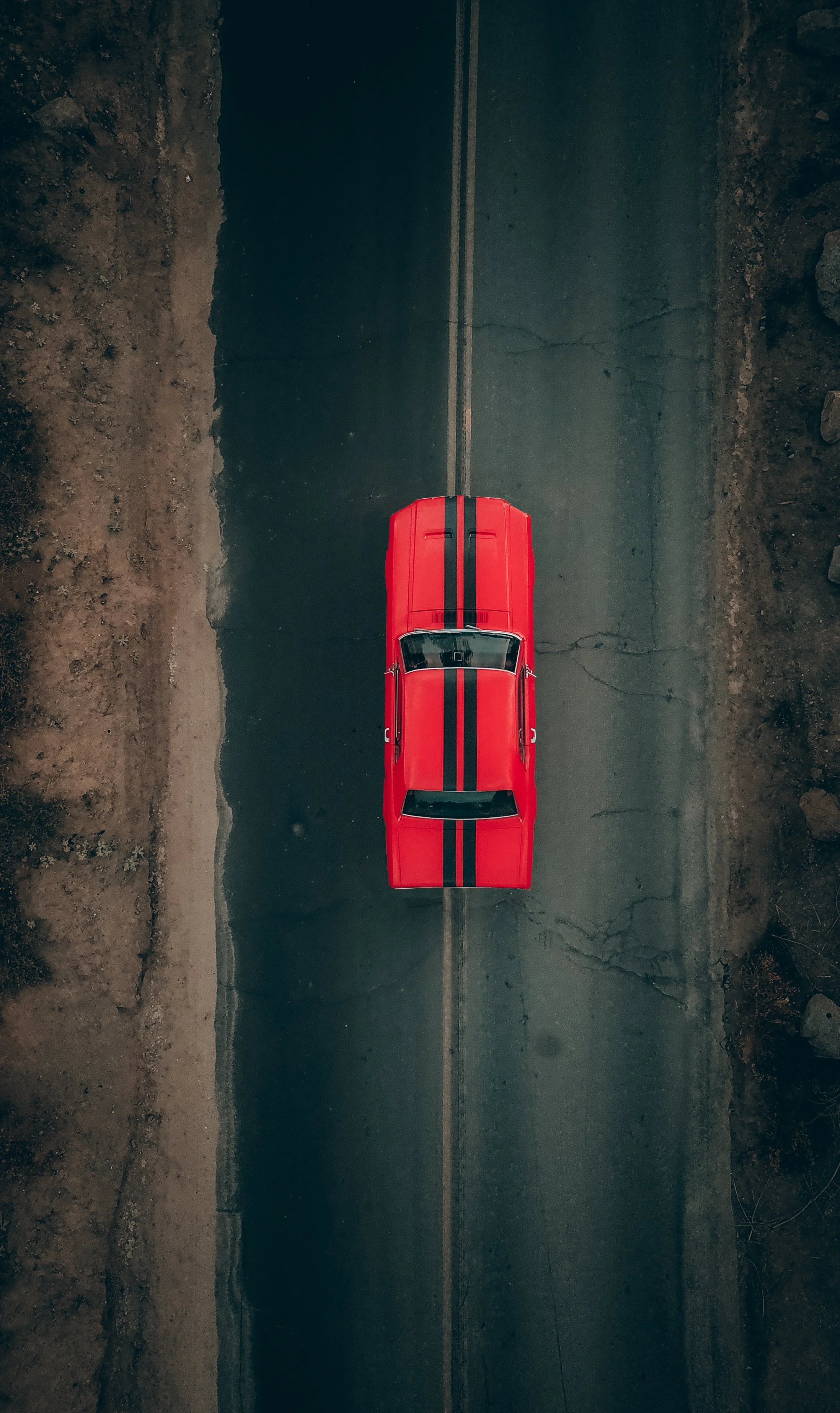 A red car with black racing stripes on an empty asphalt road, viewed from above.