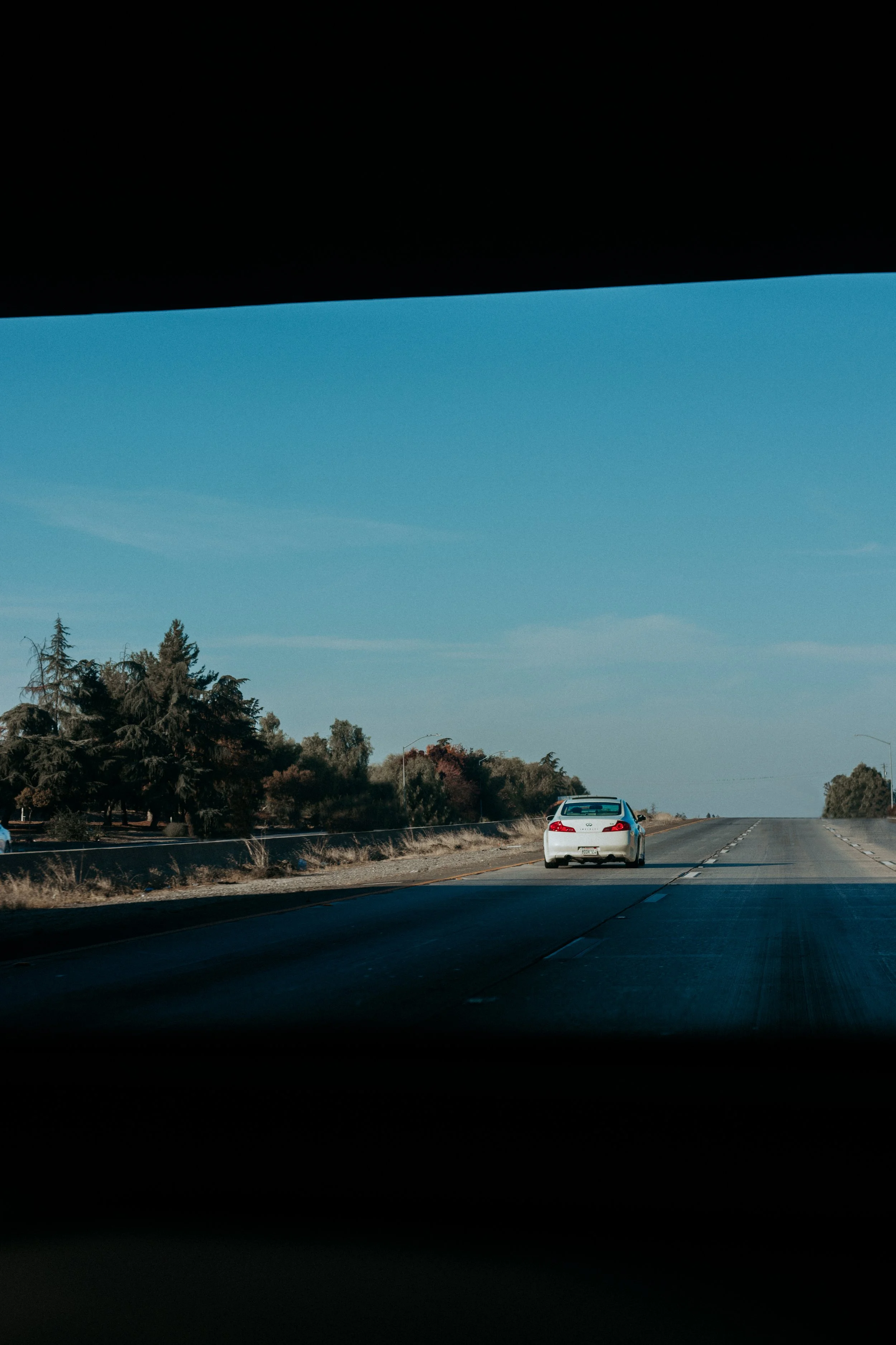 View of a highway with a white car driving along the road, surrounded by trees and open sky.
