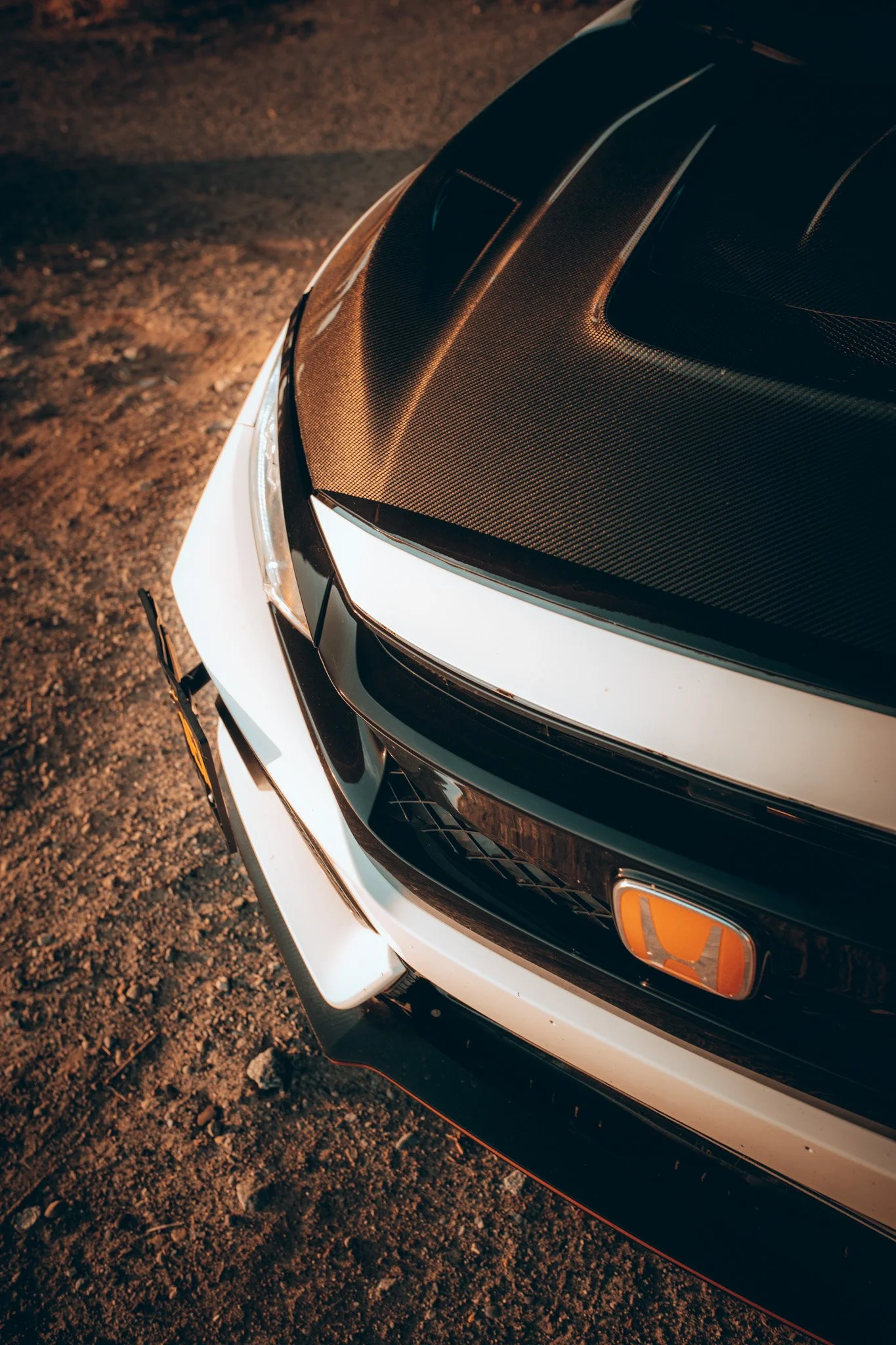 Close-up of a car's hood with a carbon fiber finish and part of the front bumper visible, parked on a gravel surface at night.