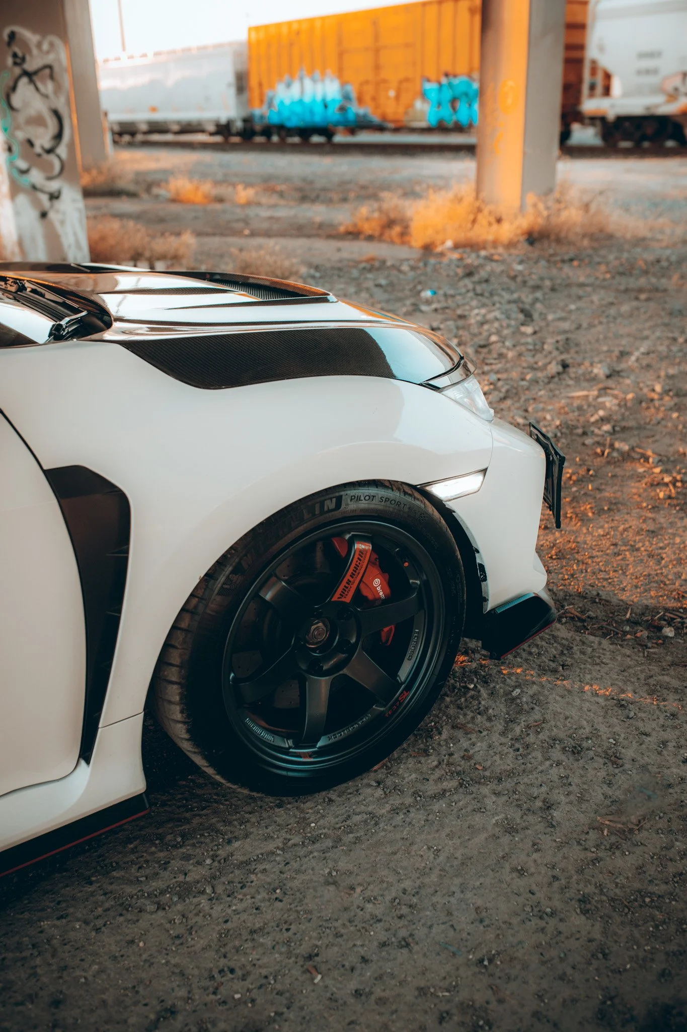 Close-up of the front part of a white sports car with black accents, parked on a dirt surface near a railway track with train cars in the background.