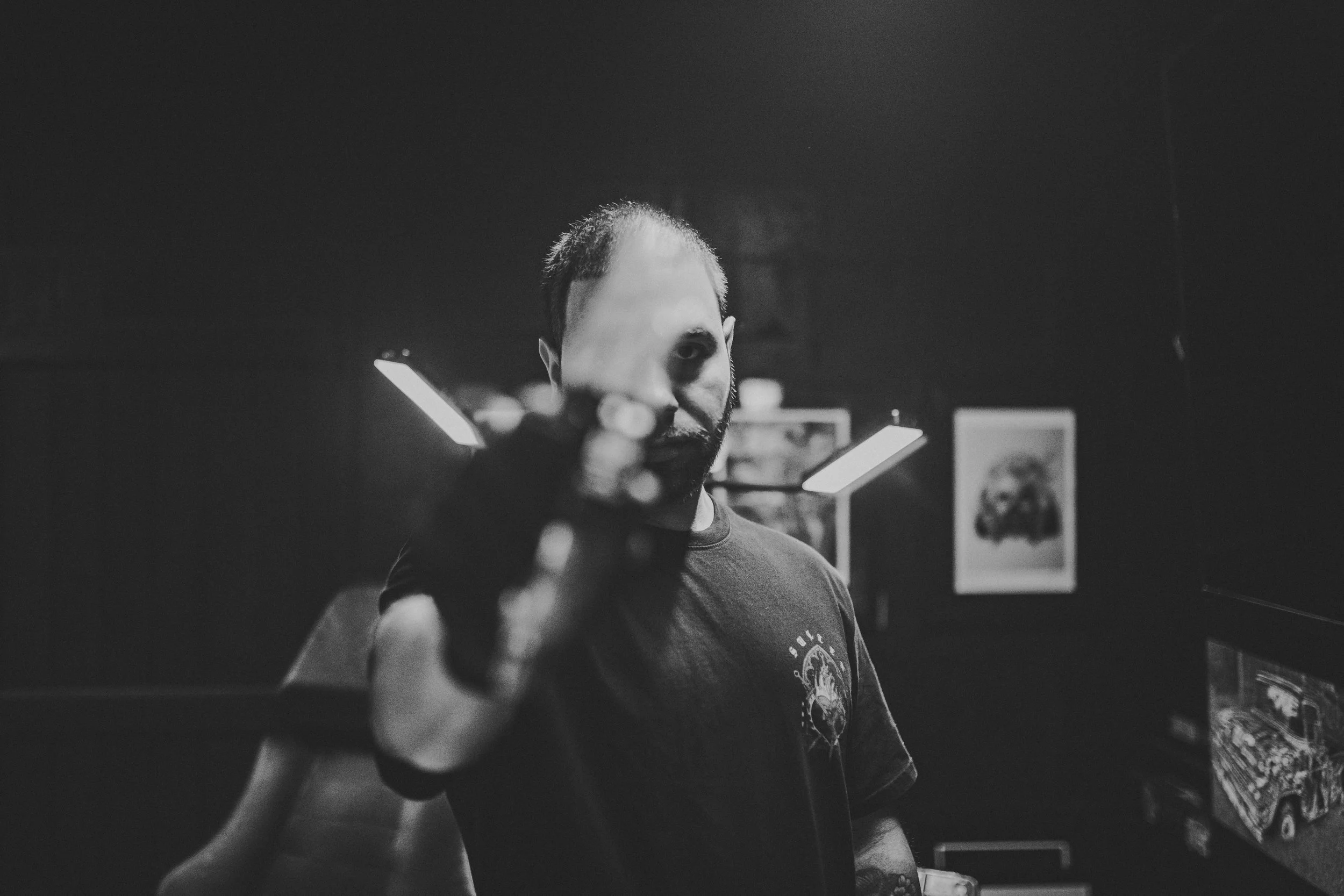 A man with short hair and a beard looking directly at the camera, holding a smartphone in front of him, in a dark room with artwork on the walls.