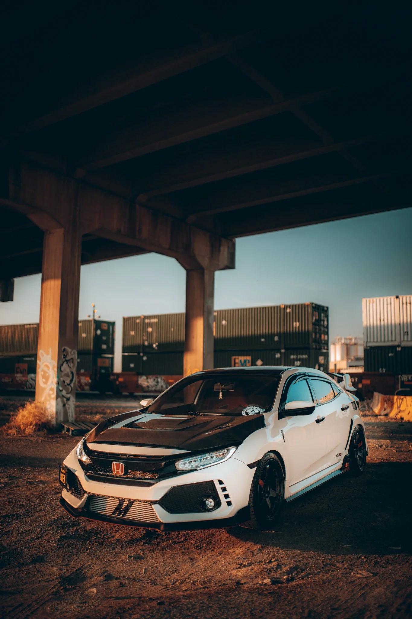 A white Honda Civic Type R with black accents parked under a highway overpass in an industrial area with shipping containers and graffiti on a pillar.