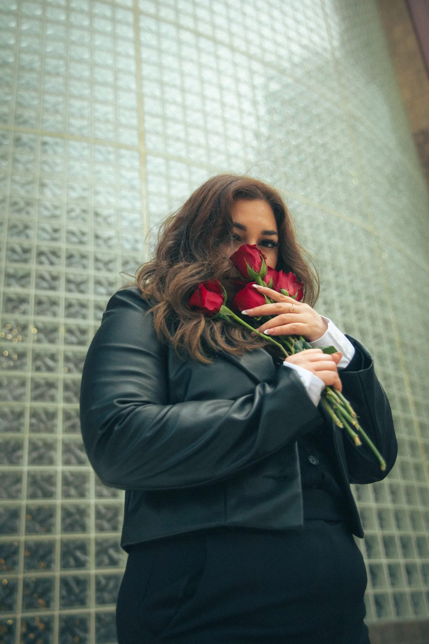 A woman with brown, curly hair holding a bouquet of red roses, standing in front of a glass block wall, wearing a black jacket and a white shirt.
