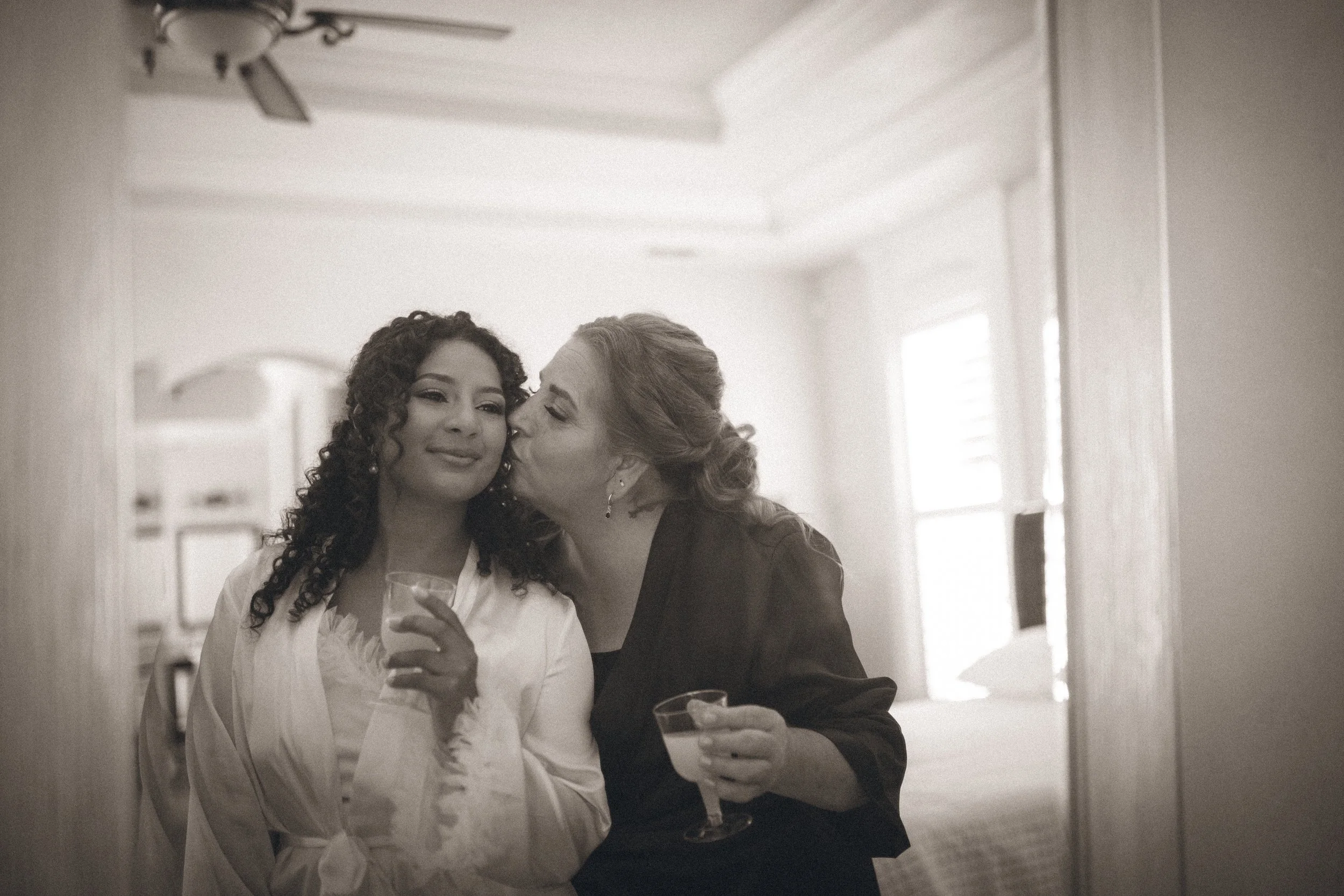 Two women sharing a kiss on the cheek at a social gathering, one holding a glass of champagne.