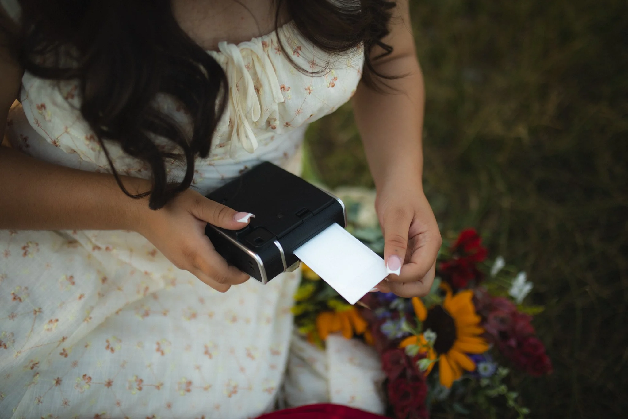 A woman in a floral dress using a portable thermal printer to print a photograph, with colorful flowers in the background.