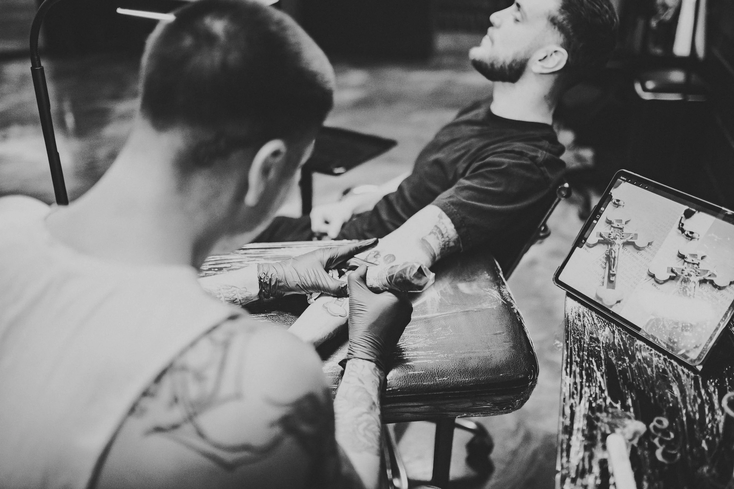 A tattooed person lying back while undergoing a tattoo procedure at a tattoo parlor, with a bartender or artist holding a tattoo machine and a tablet displaying tattoo design references.