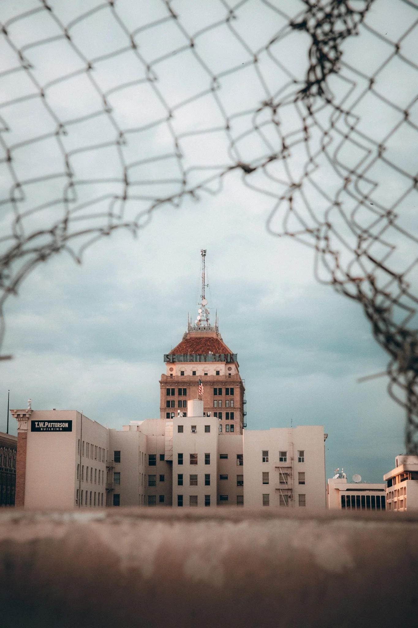 A view of multiple buildings with a tall tower in the background, seen through barbed wire in the foreground, overcast sky