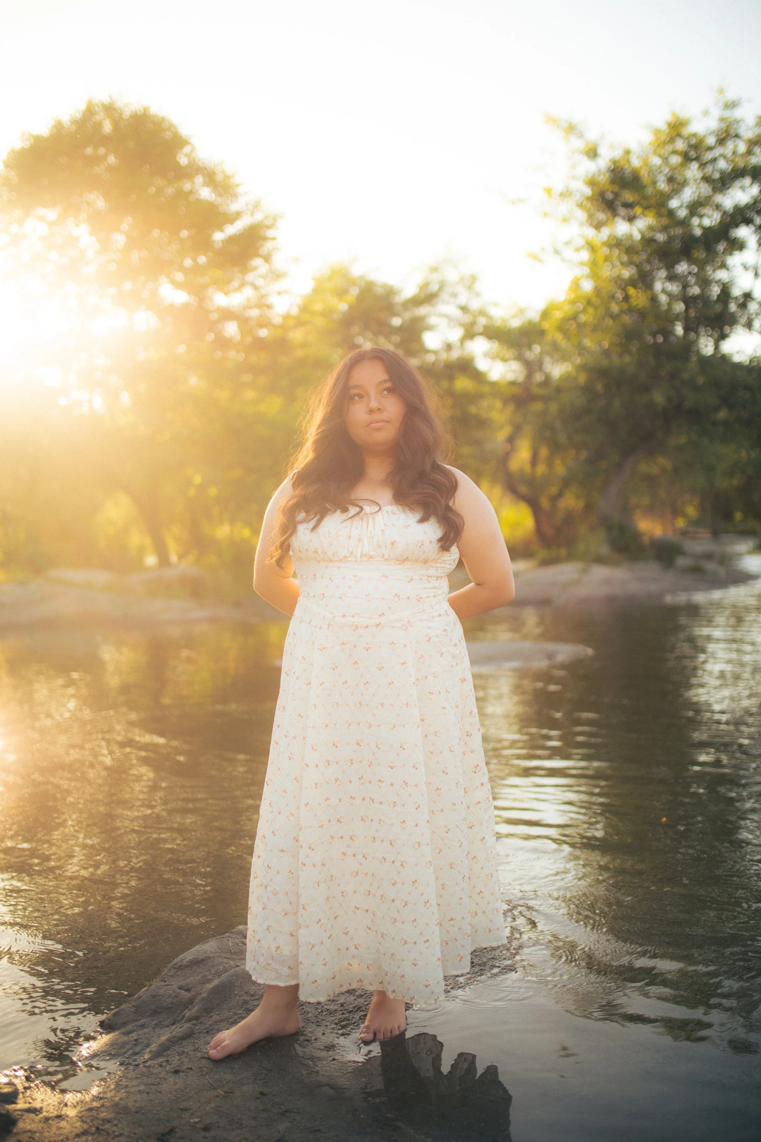 A young woman with long brown hair standing barefoot on a rock in a river during sunset, wearing a white dress with small pink floral patterns, surrounded by trees with blurry leaves in the background.