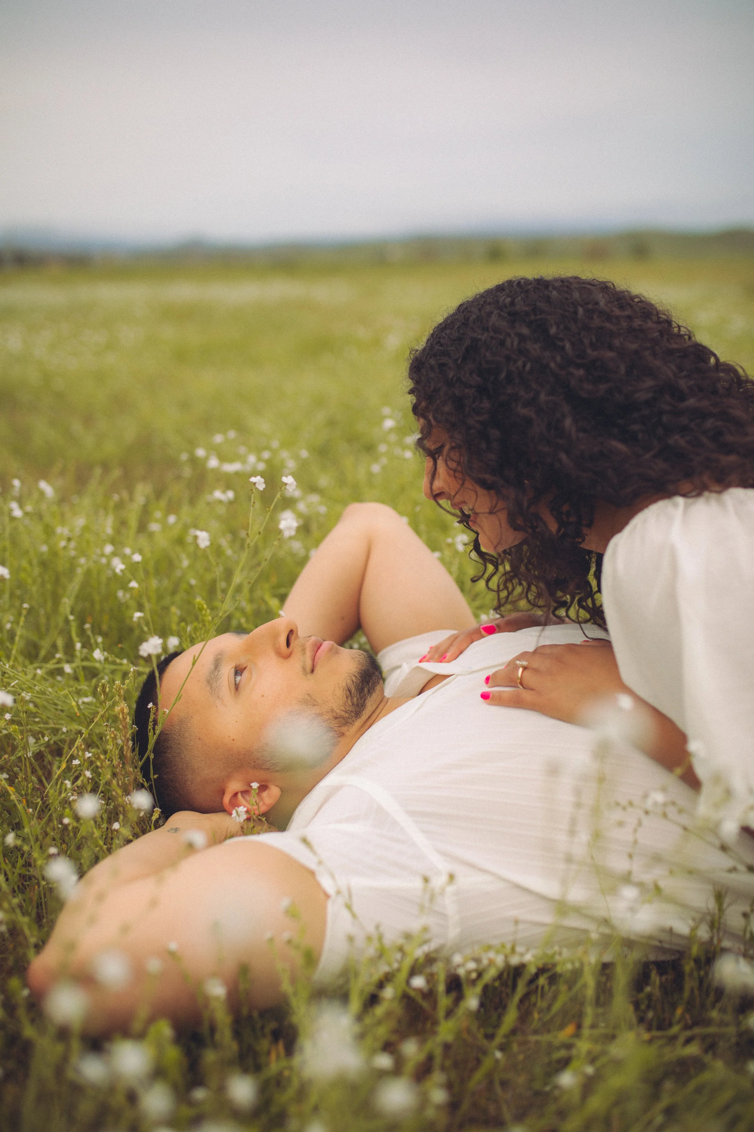 A couple has a romantic moment lying in a field of small white flowers, with the woman leaning over the man who is lying on his back, both gently touching each other.