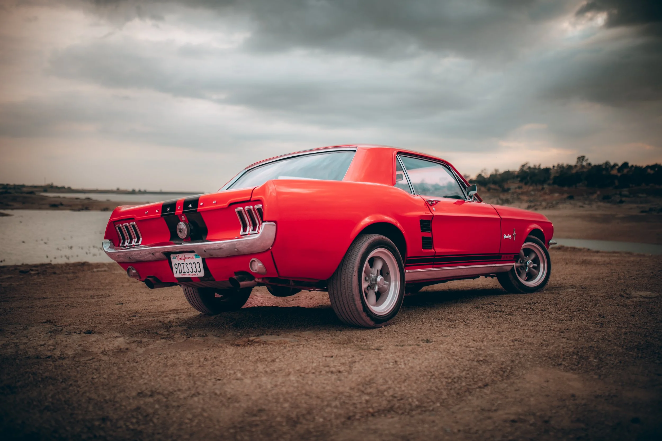 Red vintage Ford Mustang parked on a dirt surface near a body of water with cloudy sky overhead.
