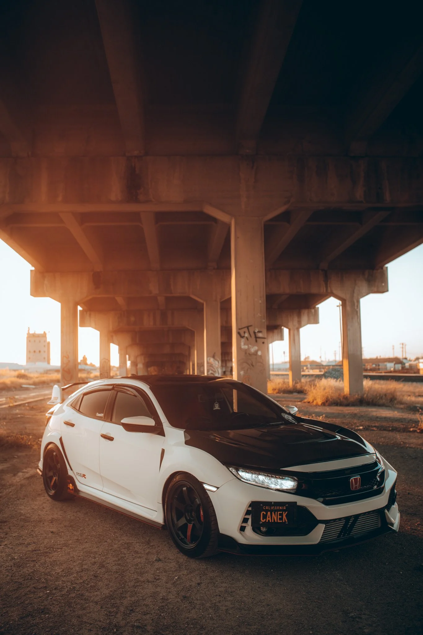 A white Honda Civic Type R parked beneath a large concrete bridge at sunset.