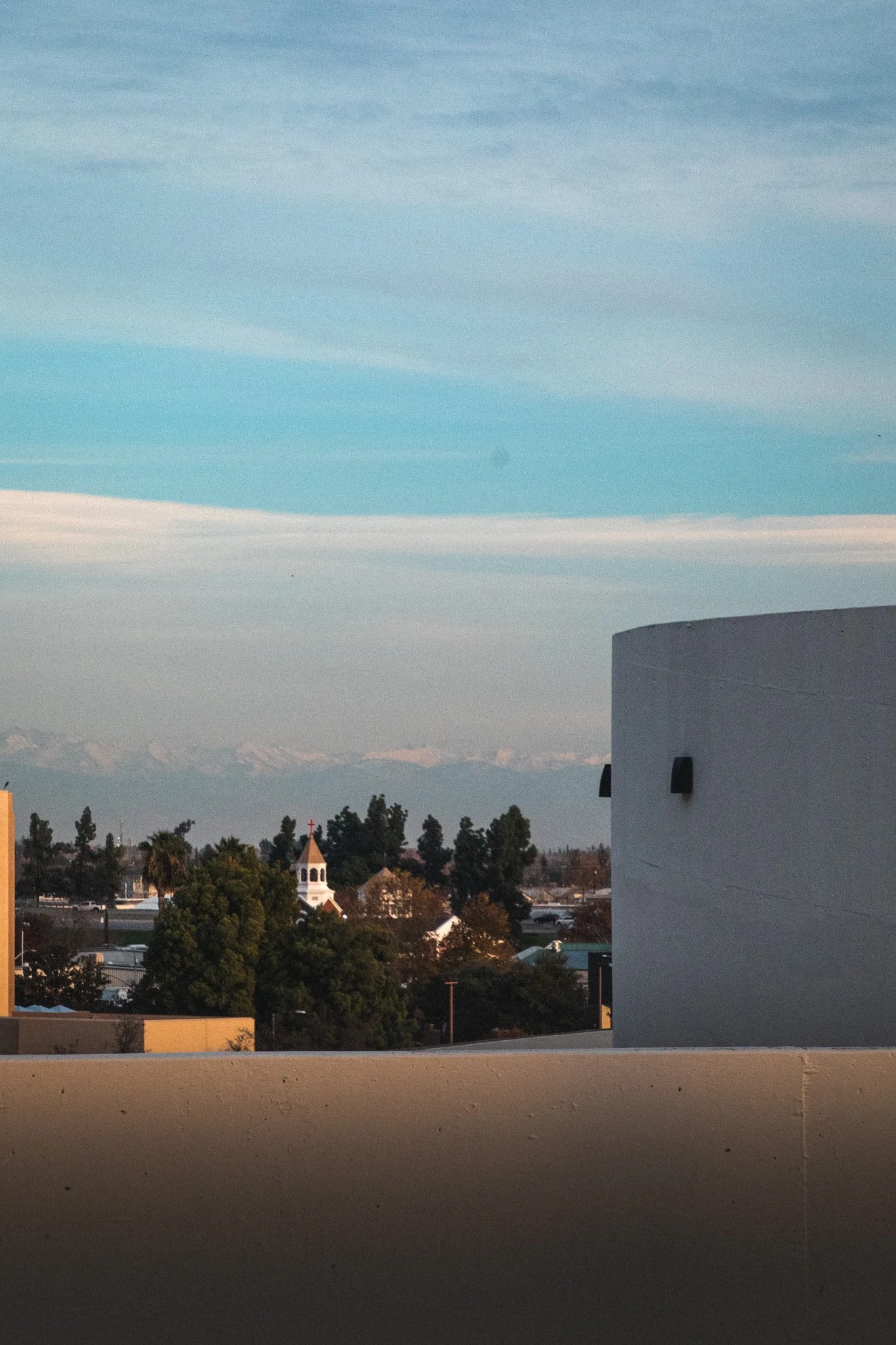 View of a small town or city with a church steeple, trees, and buildings, seen from a balcony or rooftop, with a mountain range in the background and a partly cloudy sky overhead.