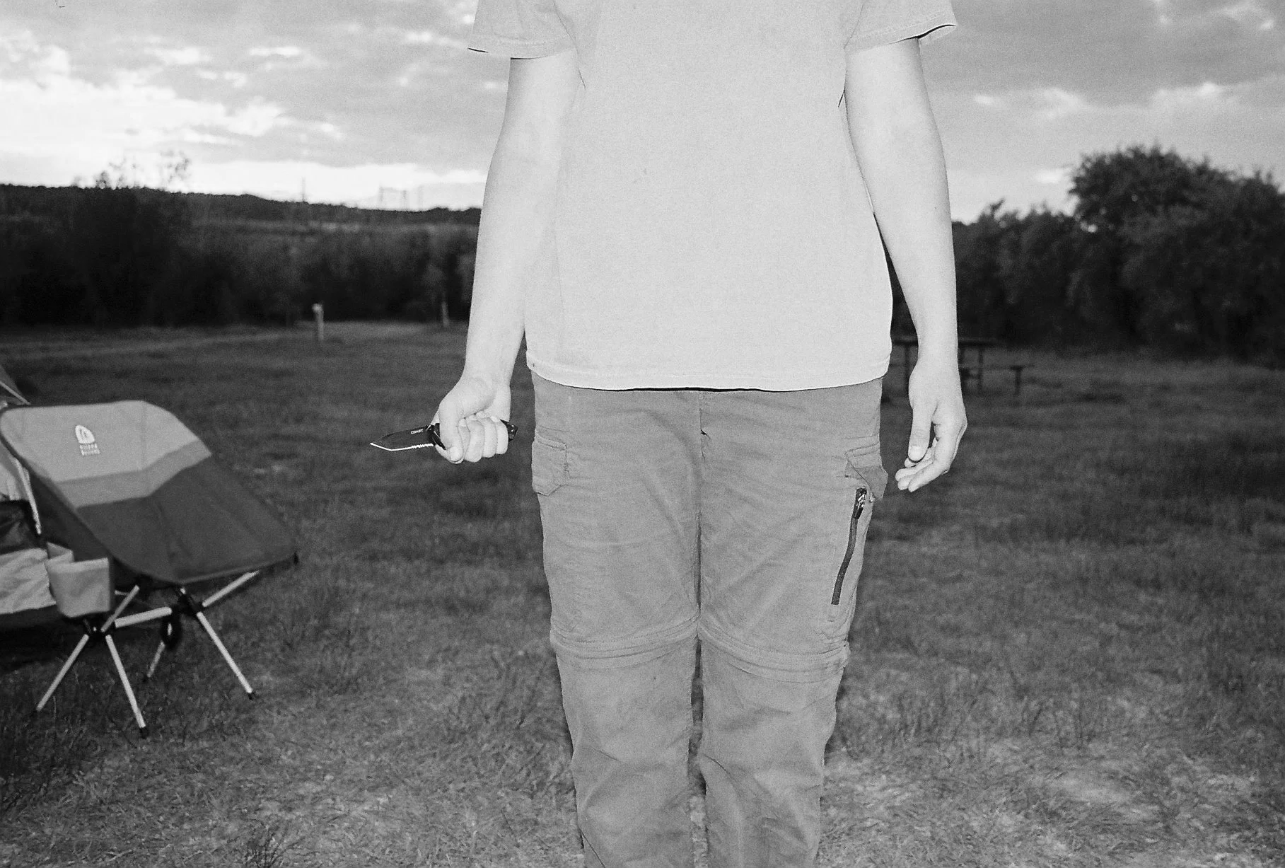 Person wearing a t-shirt and cargo shorts holding sunglasses in their right hand, standing outdoors on grass with trees and cloudy sky in the background. Camping chair is visible on the left.