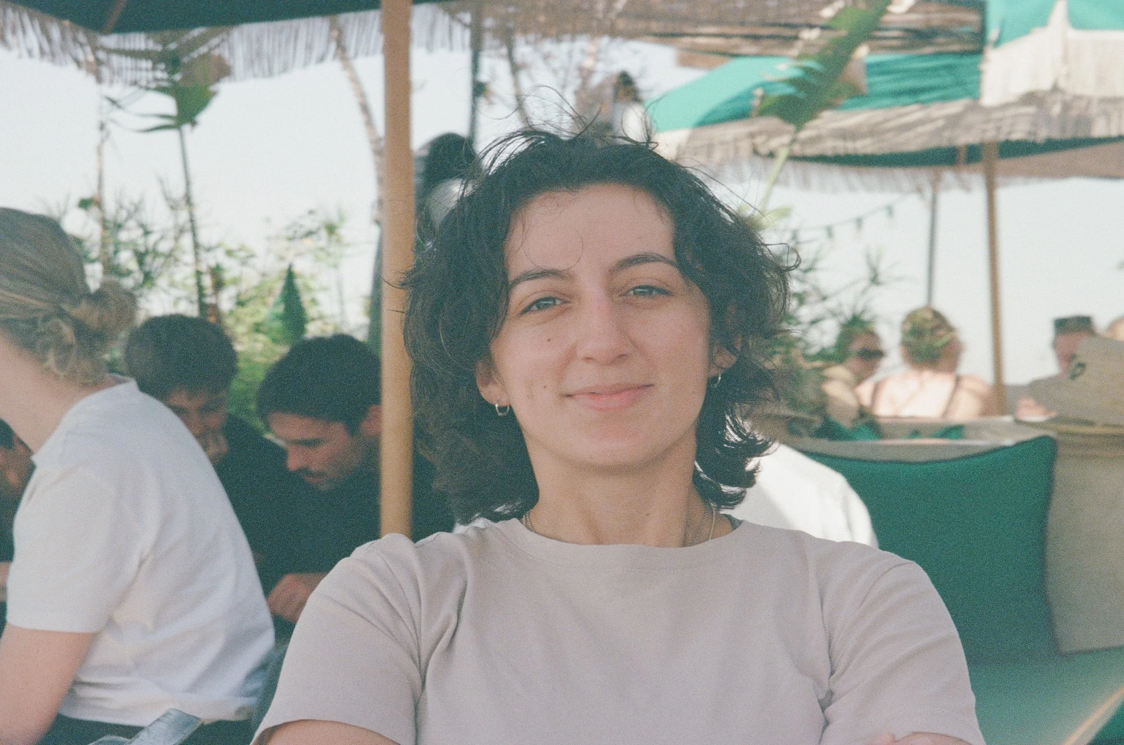 A woman with curly black hair decorated with a hair bun, wearing a white shirt, smiling slightly at the camera while taking a selfie outdoors at a gathering or event.