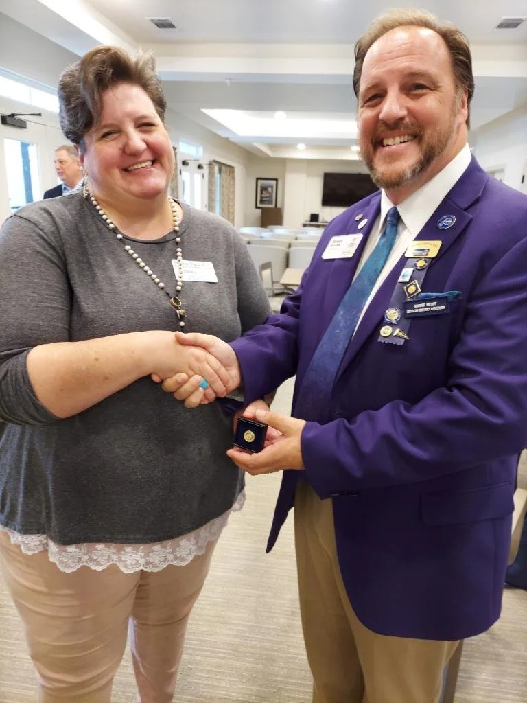 A Club Member in a gray top and beige pants shaking hands with Rotary District Governor in a purple blazer, beige pants, and a blue tie, who is giving her the Paul Harris Foundation award pin. They are smiling at an indoor event.