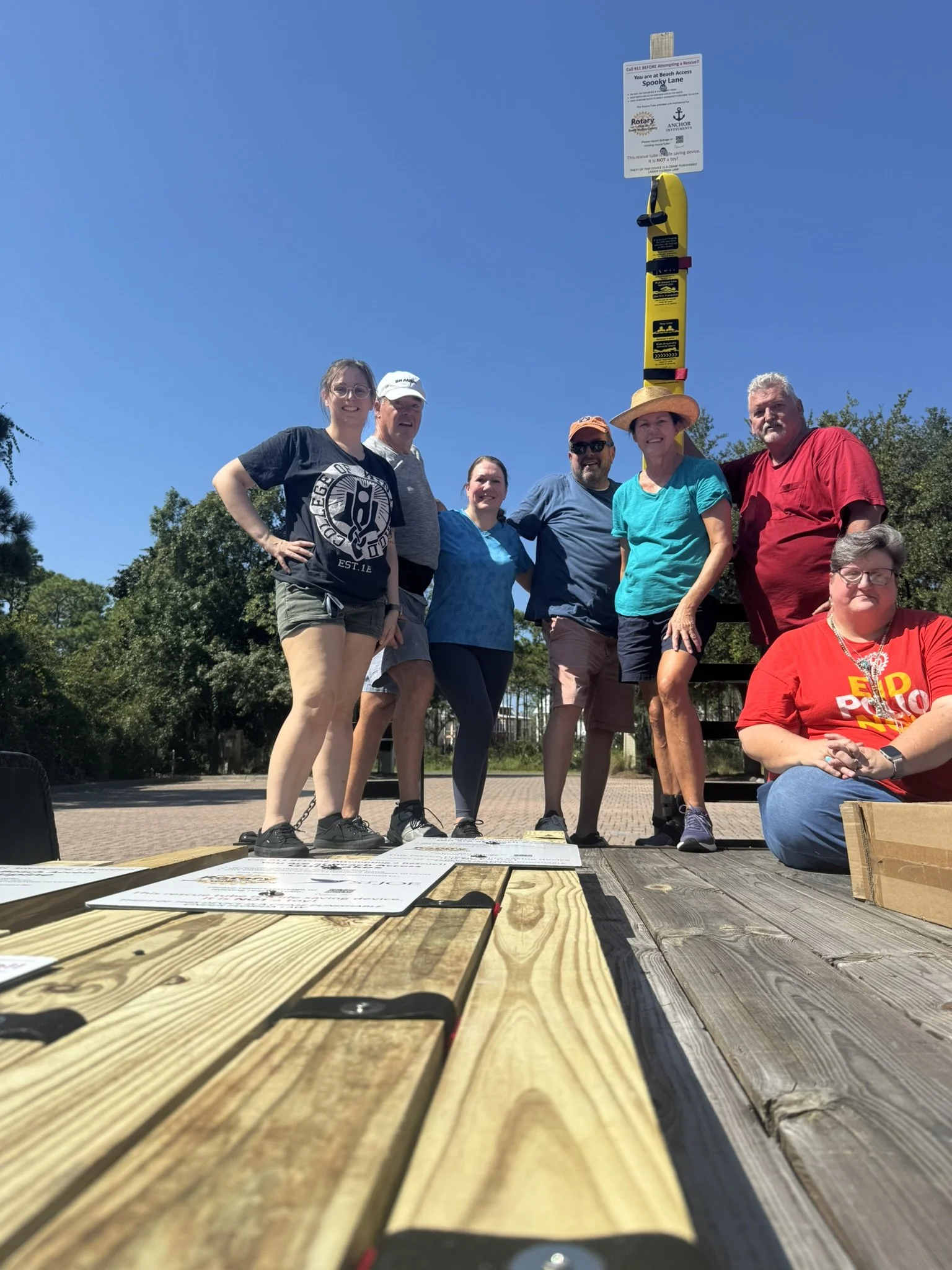 Club members posing outdoors in front of a blue sky, with a Personal Rescue Tube (PRT) sign overhead reading 'You are at Beach Access Spooky Lane,' and they are sitting on a truck trailer after assembling 10 signs for beach accesses in Walton County.