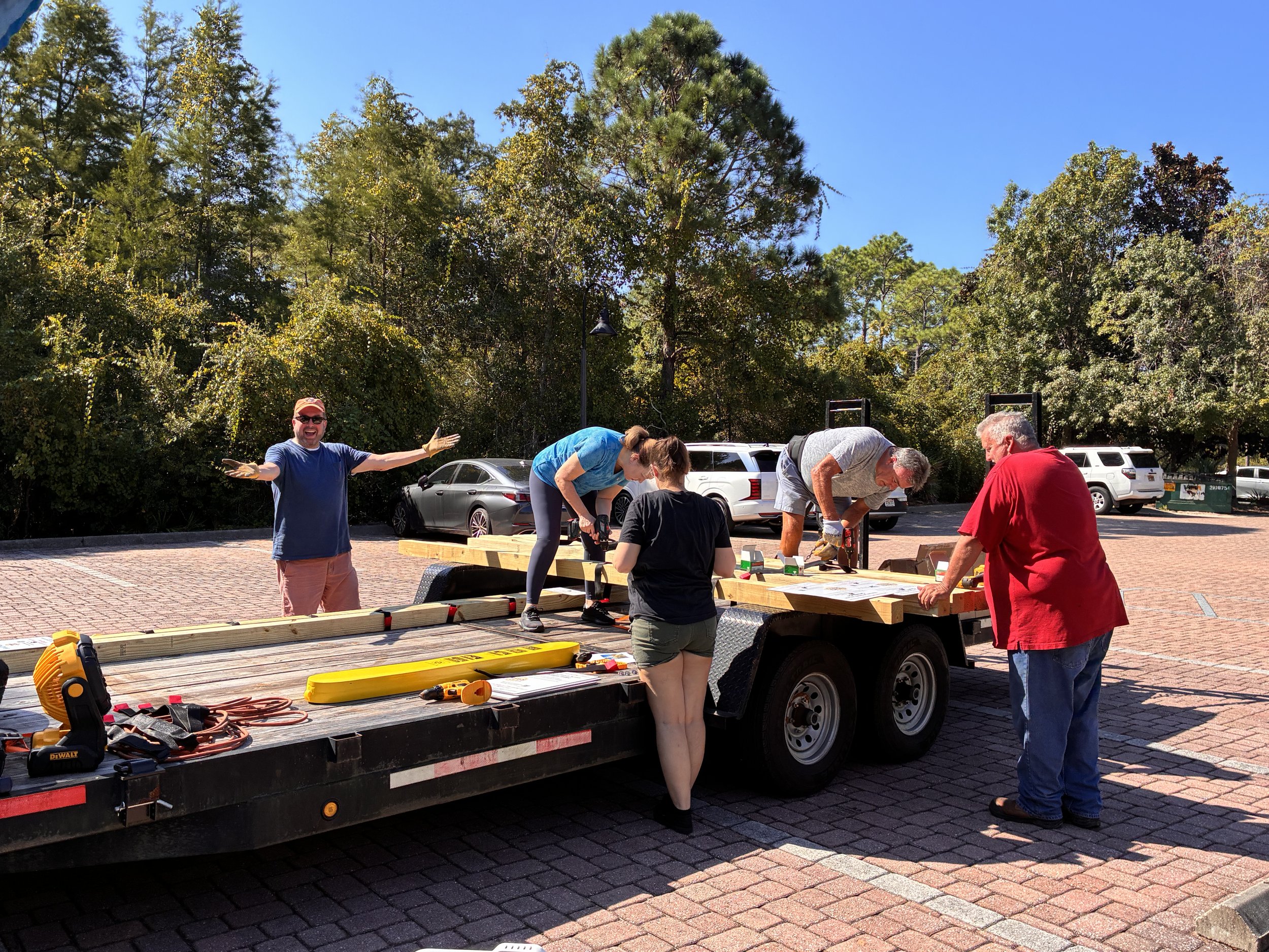 Club Members working on assembling beach signs for the Personal Rescue Tubes project on a trailer in the Dunlap & Shipman parking lot surrounded by trees and parked cars on a sunny day.