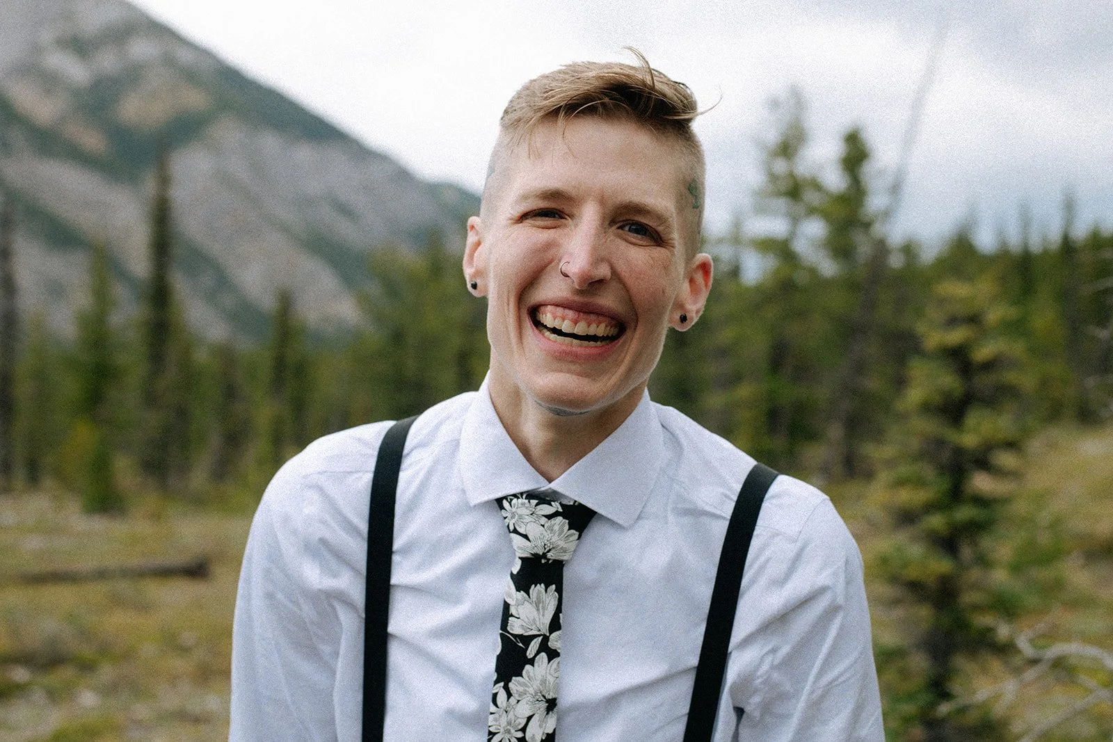 A smiling person with short hair, wearing a white shirt, floral tie, and suspenders, standing outdoors with trees and mountains in the background.