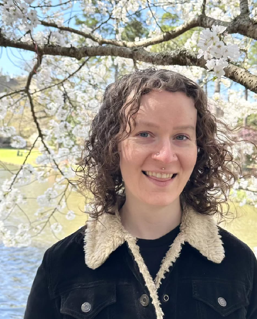 A young woman with curly brown hair, light skin, and a nose piercing, smiling outdoors under cherry blossom trees with a lake in the background.