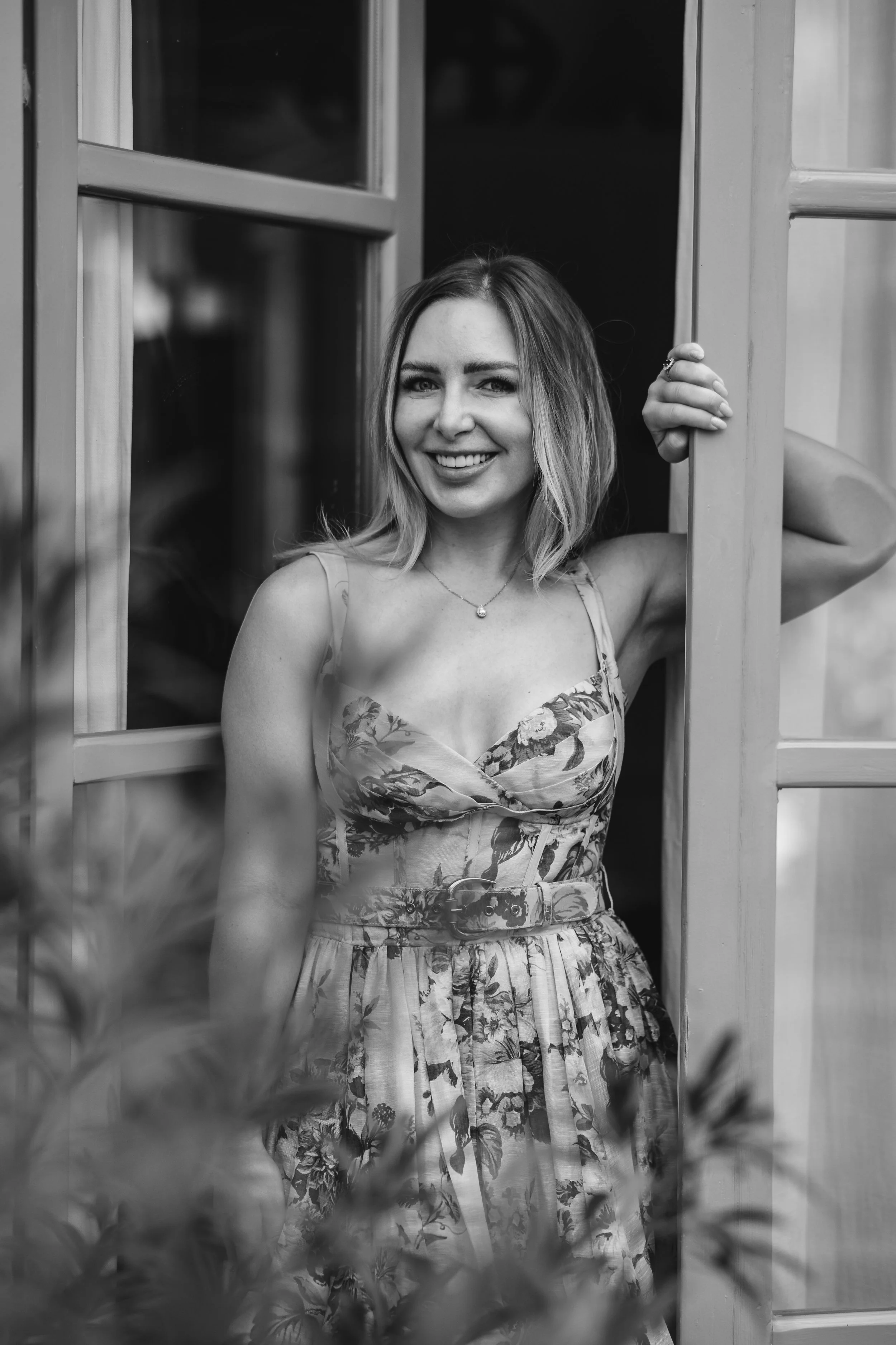 Smiling woman in floral dress peeking through a door frame, black and white photo.