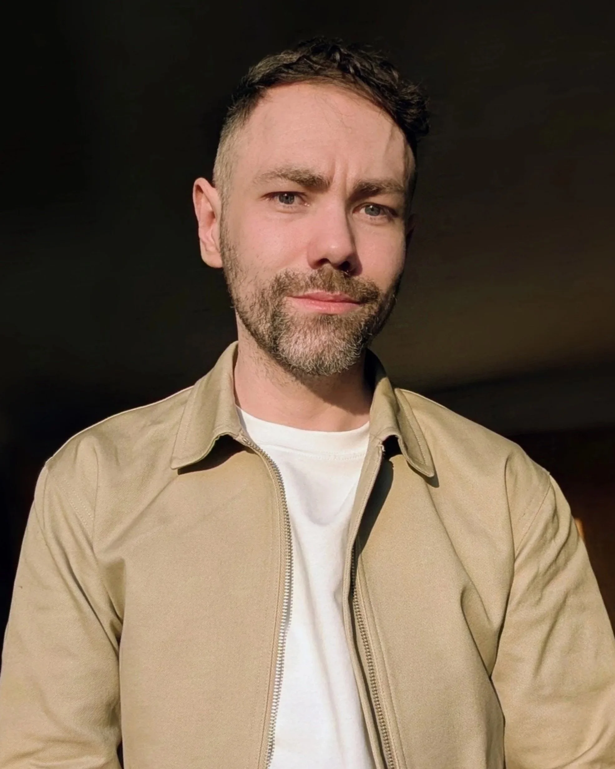 A man with short dark hair, a beard, and blue eyes, wearing a beige jacket over a white shirt, stands against a dark background.
