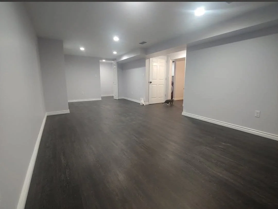 Empty living room with dark hardwood floors, white walls, and recessed ceiling lights. A small dog is near an open doorway.