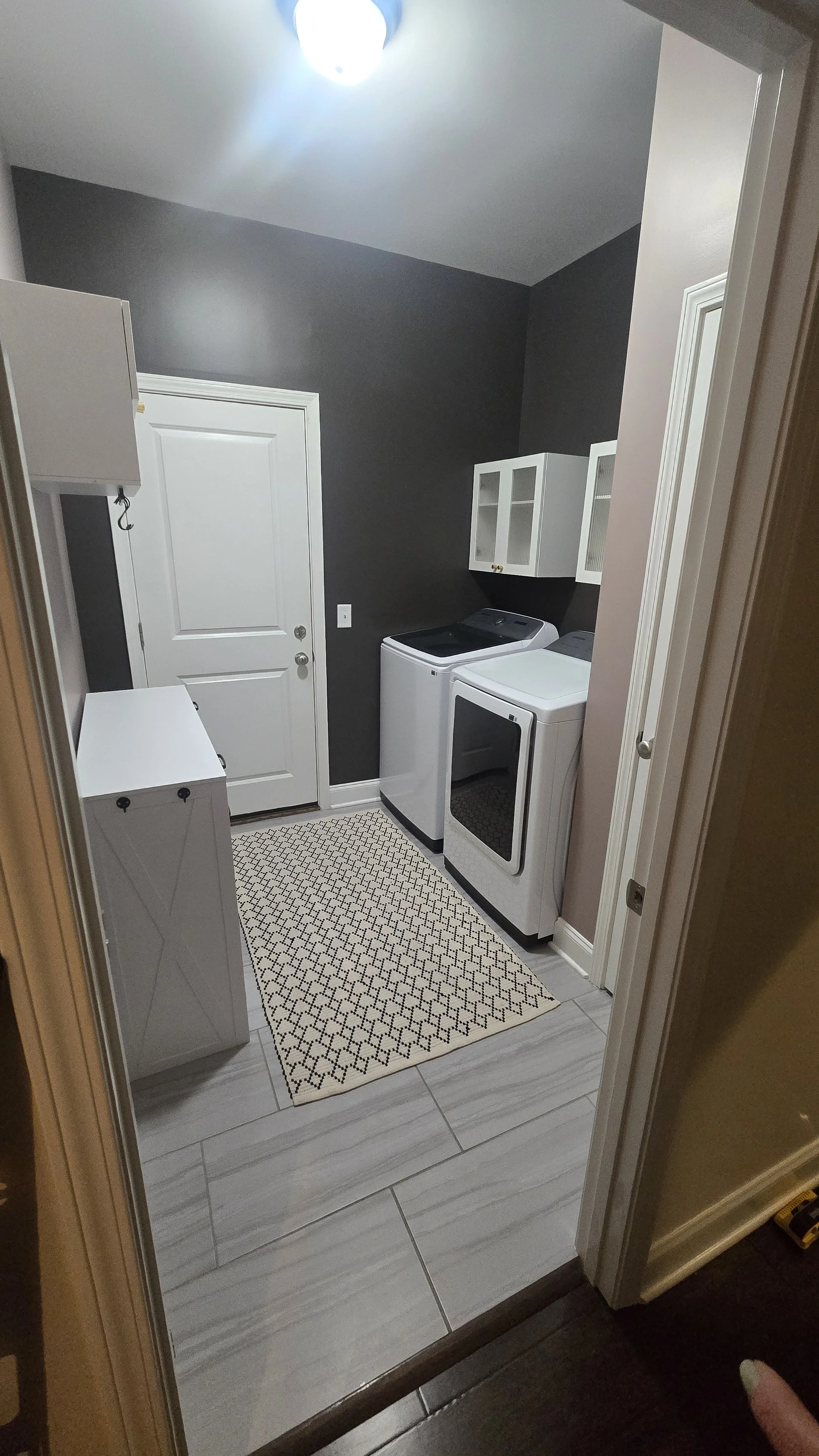 Laundry room with dark gray and beige walls, white washing machine and dryer, white wall-mounted cabinet, small white chest, patterned rug, white door, and tiled floor.
