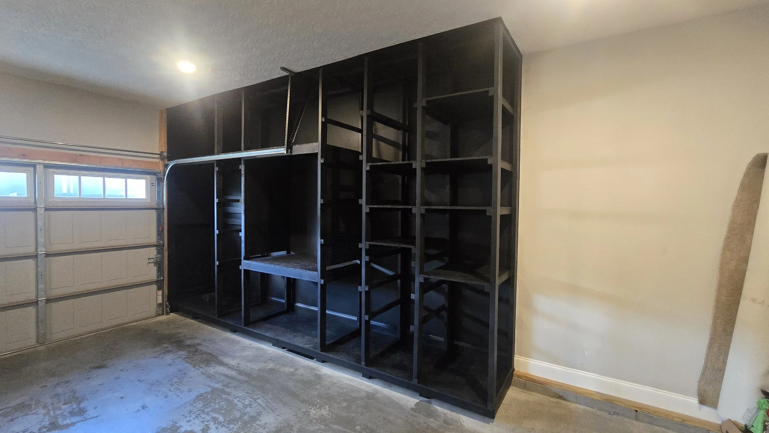 Unfinished garage with a large black shelving unit along the wall, next to a garage door and a beige wall.