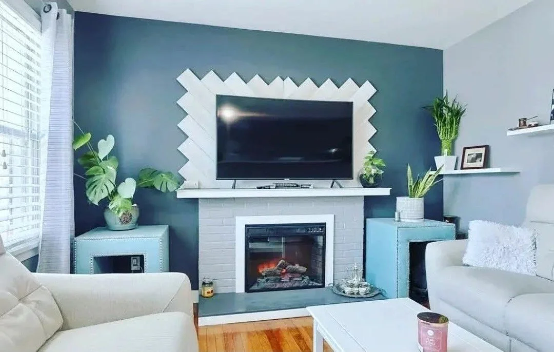 Living room with a dark blue accent wall, a white brick fireplace, a flat-screen TV above the fireplace, and green houseplants on blue and white furniture.