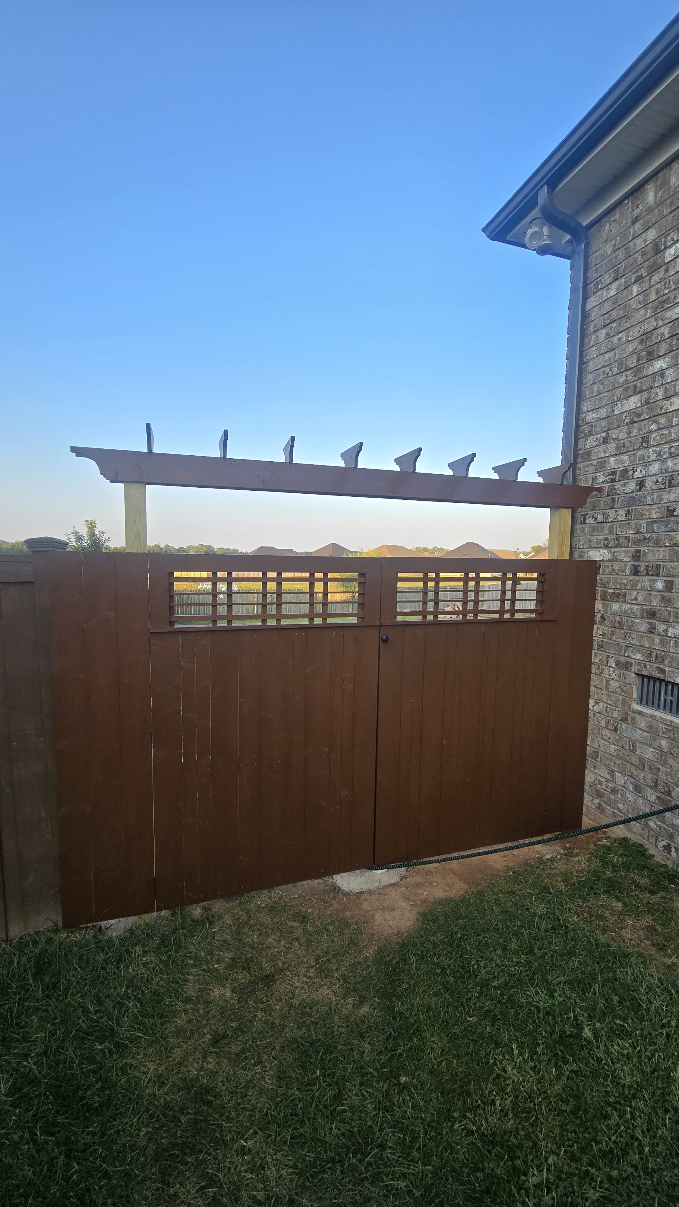 A wooden gate with a lattice design at the top, installed in a backyard next to a brick house, with a wooden frame structure above it and a blue sky in the background.