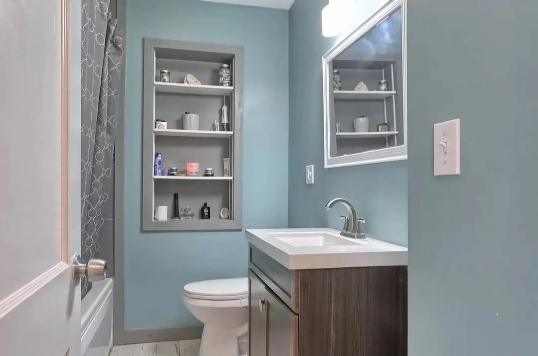 Small bathroom with a toilet, a dark wood vanity with a white sink, a mirror, a built-in wall shelf with toiletries, light blue walls, a shower with a curtain, and a window with patterned curtains.