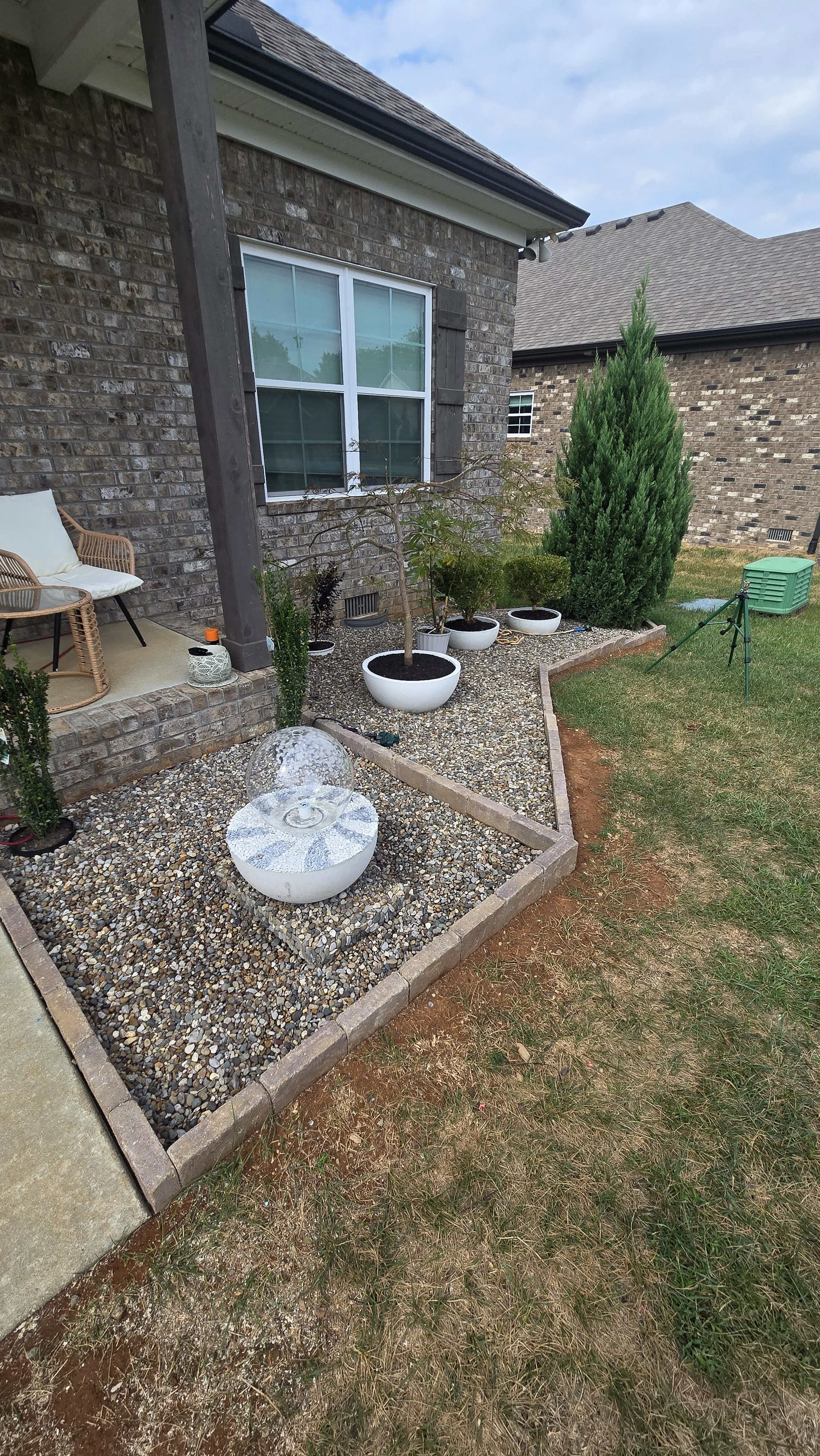 Backyard garden with potted plants, including a small tree, and a decorative water fountain on gravel next to a brick house.