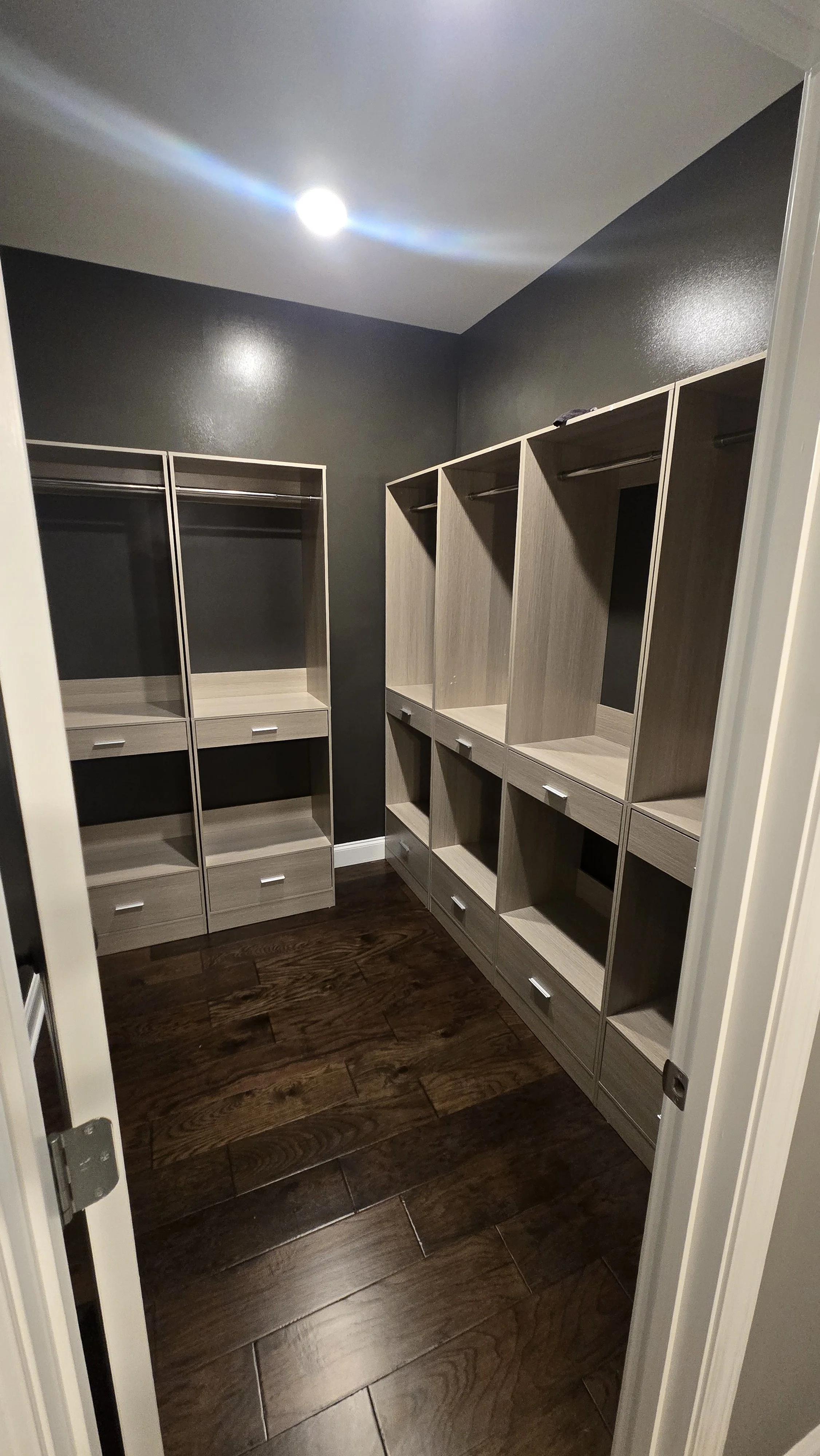 Empty walk-in closet with built-in shelves and drawers, dark wooden flooring, and dark gray painted walls, illuminated by a ceiling light.
