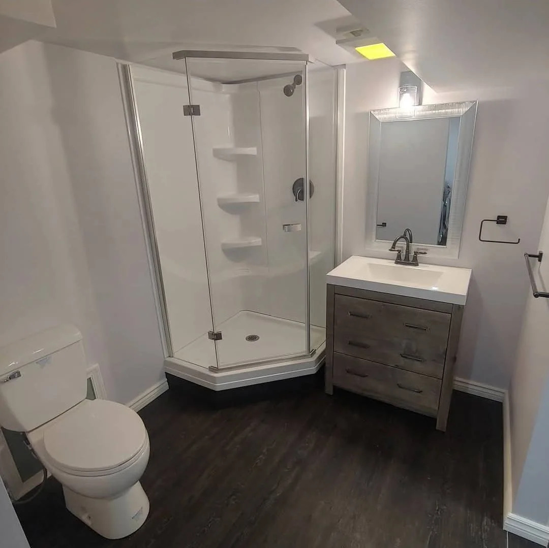 A small bathroom with a white toilet, a corner shower with glass doors, a gray vanity with a sink, a mirror above the vanity, and dark wood flooring.