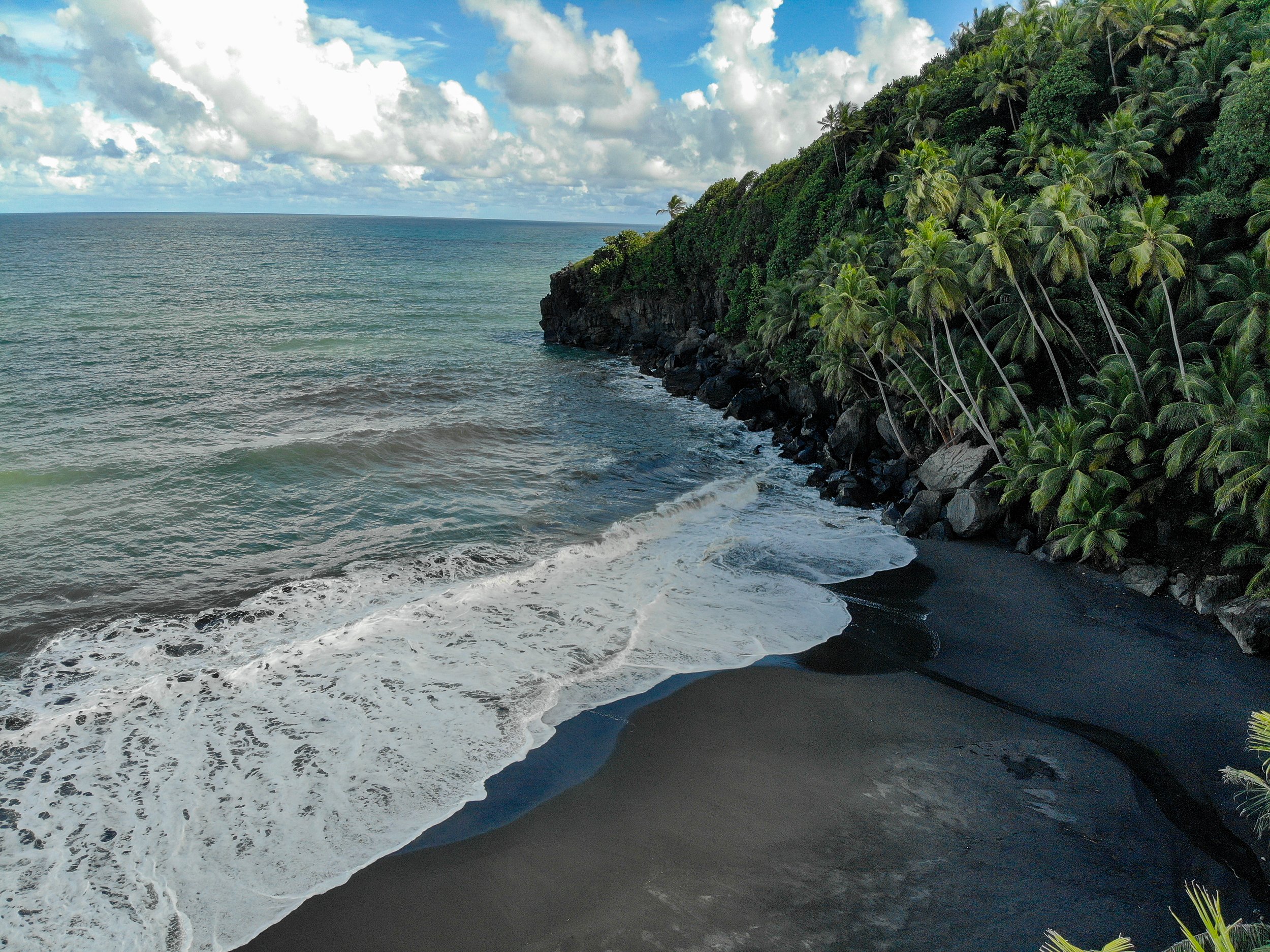 A scenic view of a black sand beach with white waves crashing on the shore, a lush green hillside with palm trees, and a partly cloudy sky. This is located at the Black Point Tunnel Heritage Park in St Vincent