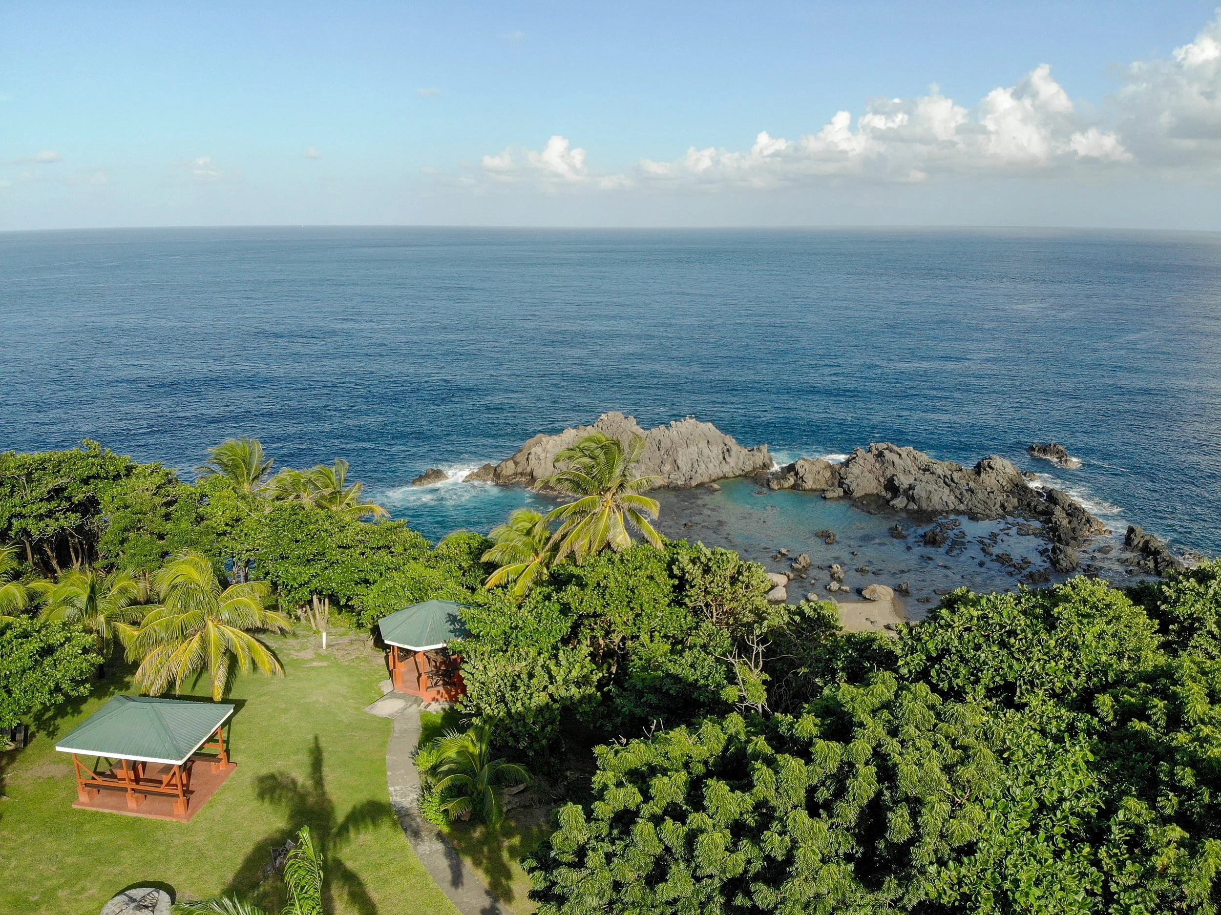 A lush green area with trees and a small building with a gazebo, near a rocky coastline and the sea in the background with partly cloudy skies. This is located at Owia Heritage Park in St Vincent.