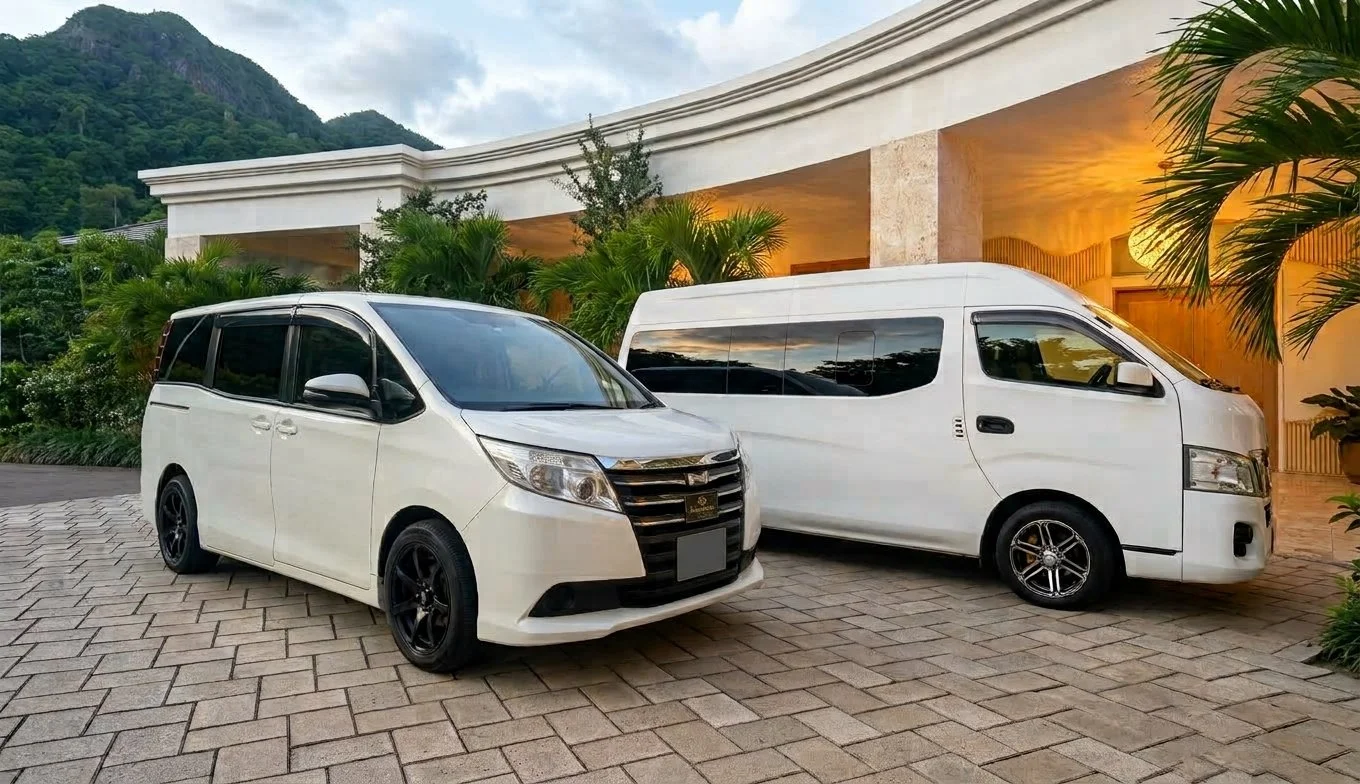 Two white vans parked on a driveway in front of a house with tropical plants and a mountain in the background. These are the Fleet Vehicles of Barry's Island Transfers