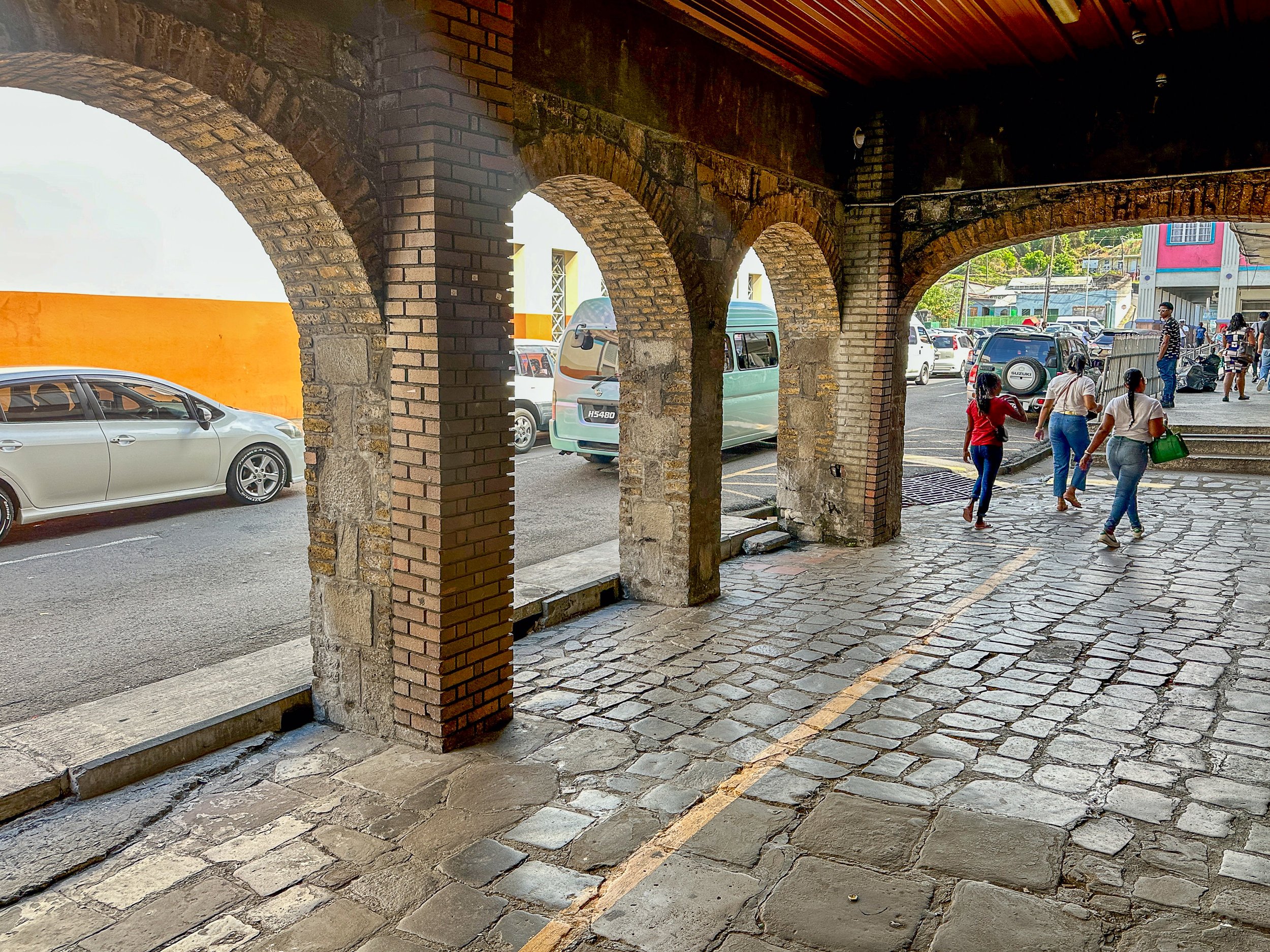 A view of a street scene taken from under a brick archway with multiple arches. Vehicles including cars and a small van are parked along the street. Pedestrians walk past the parked cars, some carrying bags. Buildings with colorful facades and greenery are visible in the background.