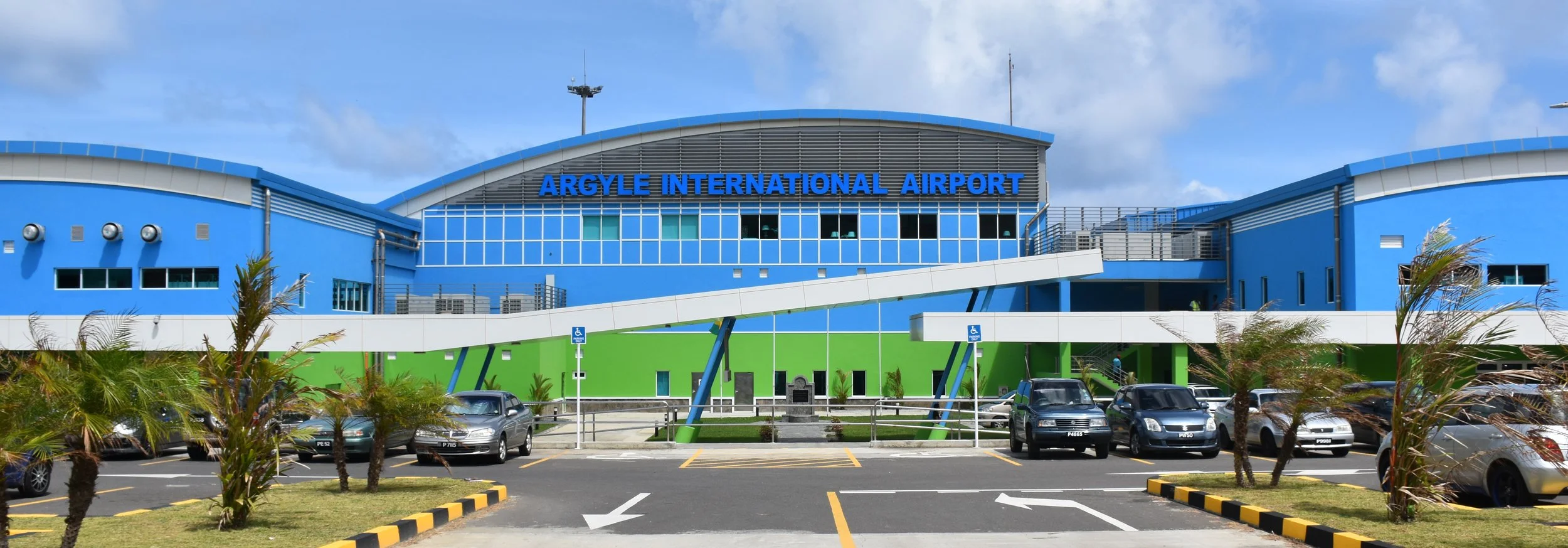 Front view of Argyle International Airport building with blue and green exterior, parking lot with cars, palm trees, and a partly cloudy sky.